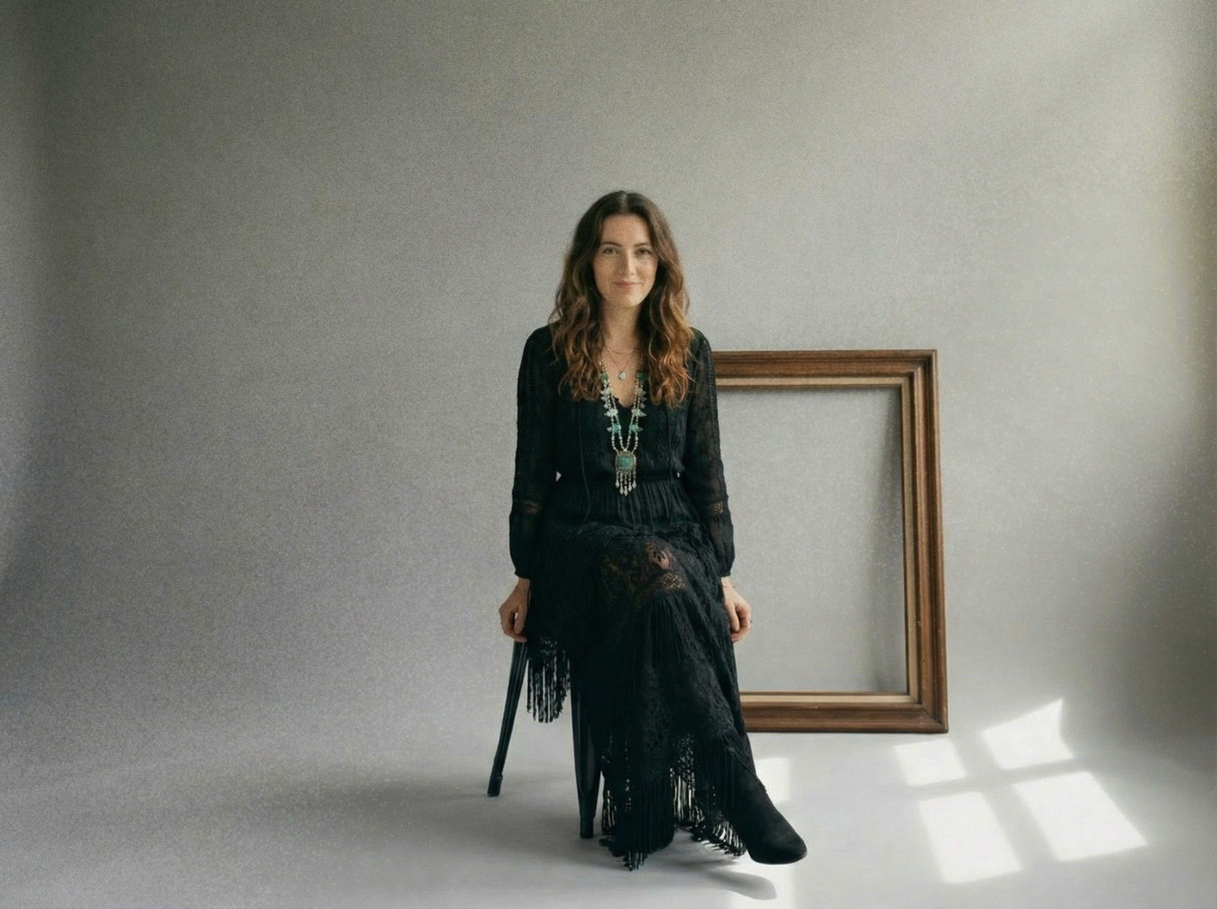 A woman in a black lace dress and boho jewelry sits on a black chair in a minimal studio with a blank wall. An empty wooden picture frame lies on the floor behind her, and sunlight creates a pattern of squares on the floor.