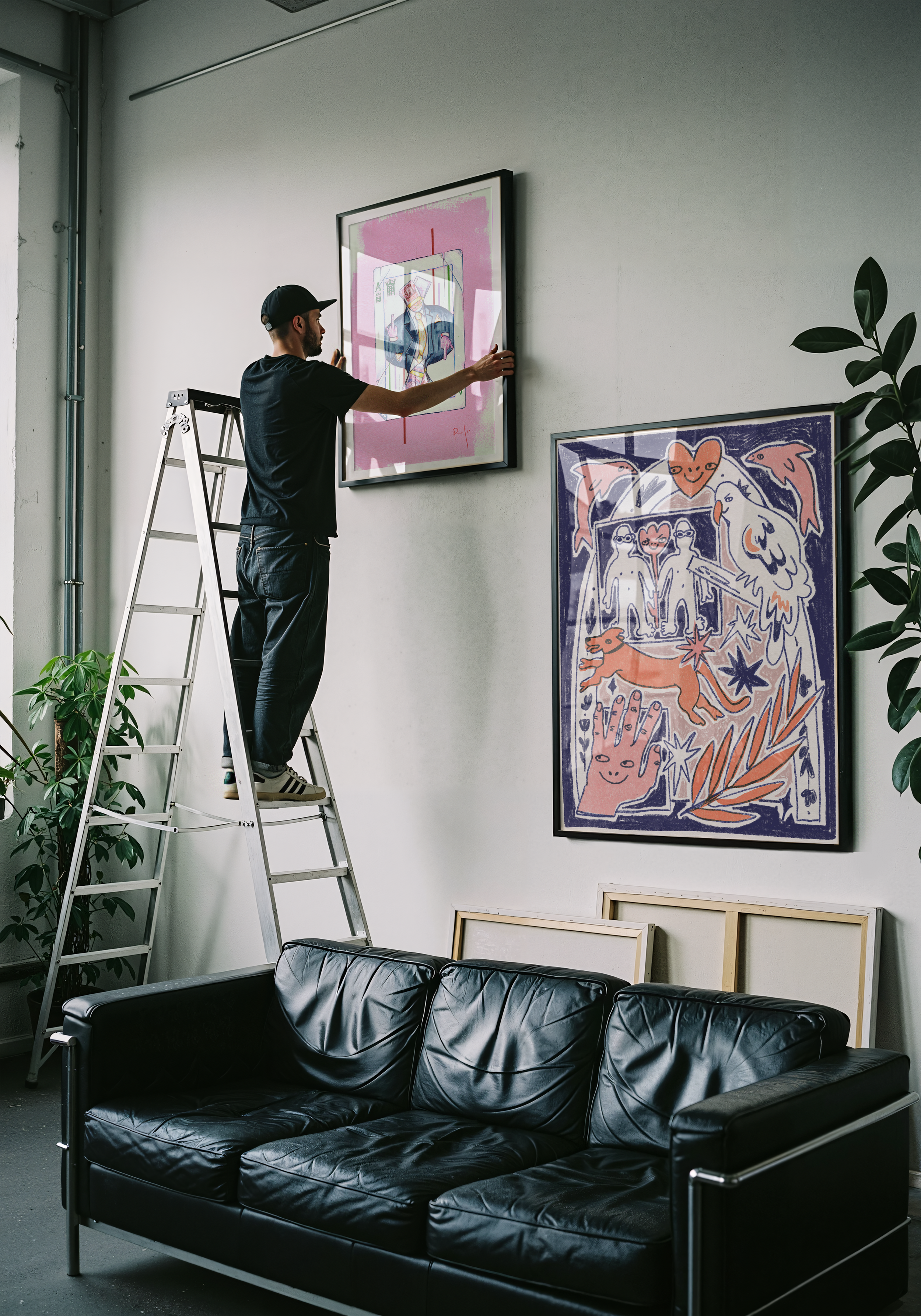 A man on a ladder hanging a framed artwork on a white wall in a modern art gallery or studio.
