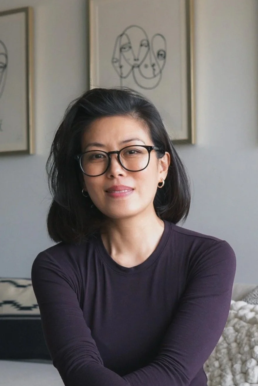A woman with black hair, glasses, and hoop earrings, wearing a dark long-sleeve shirt, is sitting in a room with abstract line art pictures on the wall behind her.