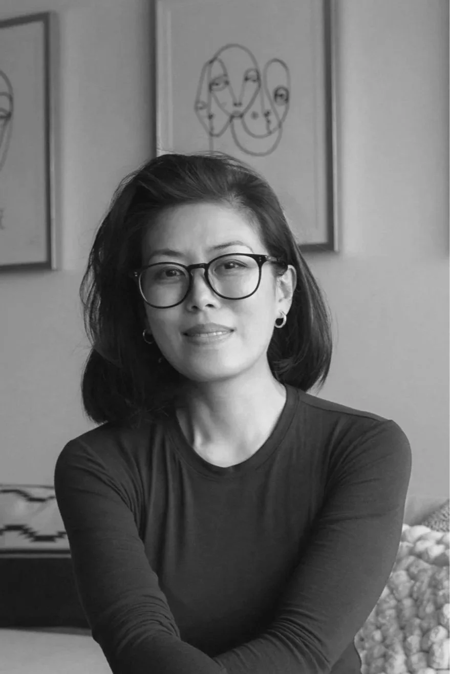 A woman with glasses and earrings, smiling softly, sitting indoors with abstract line art on the wall behind her.