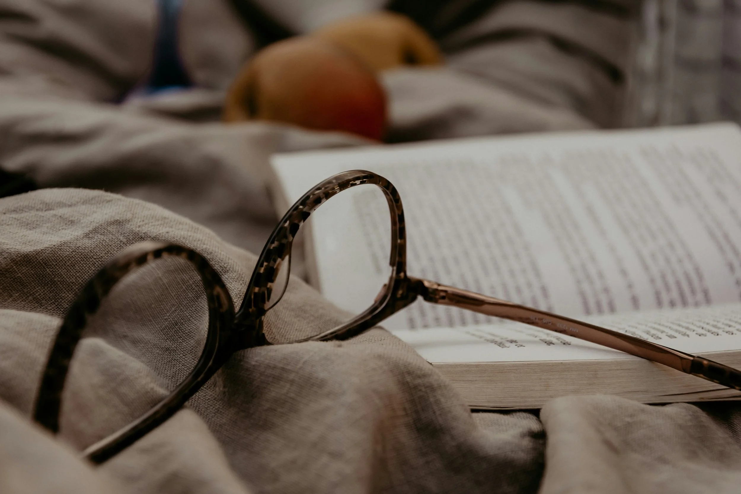 A close-up of glasses with a tortoiseshell pattern resting on a book or magazine with text, surrounded by a beige cloth and a soft fabric background, with a blurred yellow object in the background.