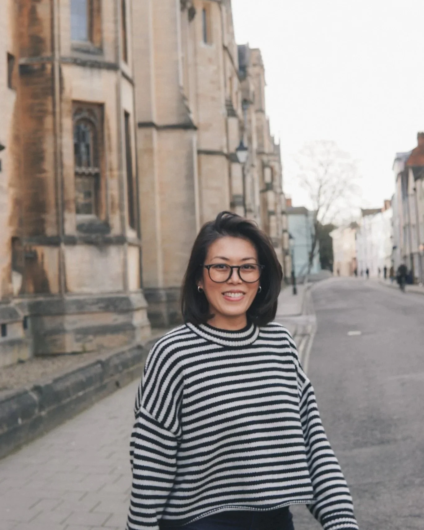 A woman with glasses and shoulder-length dark hair smiling, standing on a city street with historic stone buildings and leafless trees in the background.