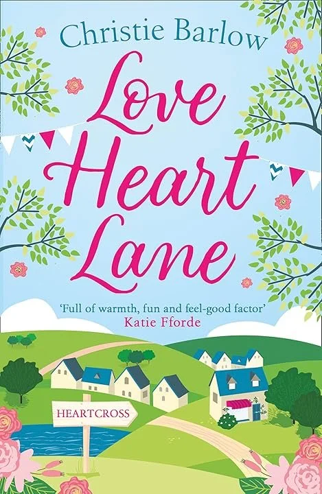 Book cover titled "Love Heart Lane" by Christie Barlow. Features a colourful, whimsical illustration of a village with houses, trees, and a small pond labeled "HEARTCROSS," with decorative flowers and a blue sky.