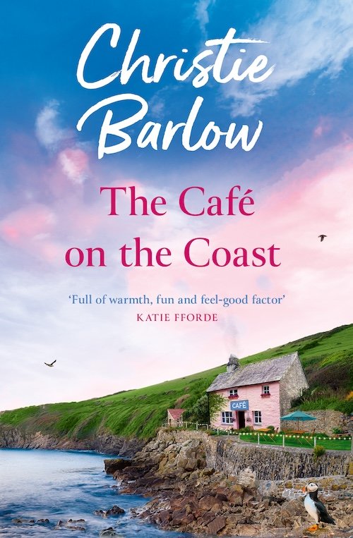Book cover for 'The Café on the Coast' by Christie Barlow. The cover features a scenic coastal landscape with a pink cottage next to the water, green hills, and a sky with pink and blue clouds.