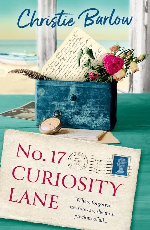 Book cover for "No. 17 Curiosity Lane" with a subtitle about forgotten treasures, a blue box containing a letter and a bouquet of pink and white roses, a pocket watch, and a postcard on a table with a beach scene in the background.