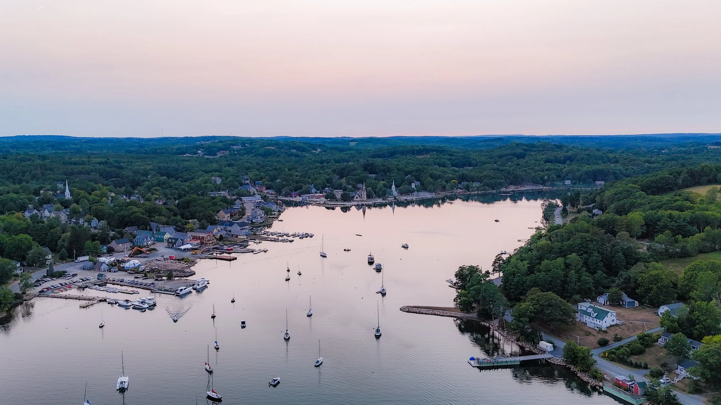Aerial view of a coastal town with boats on a calm river, surrounded by lush green trees and houses, at sunset.