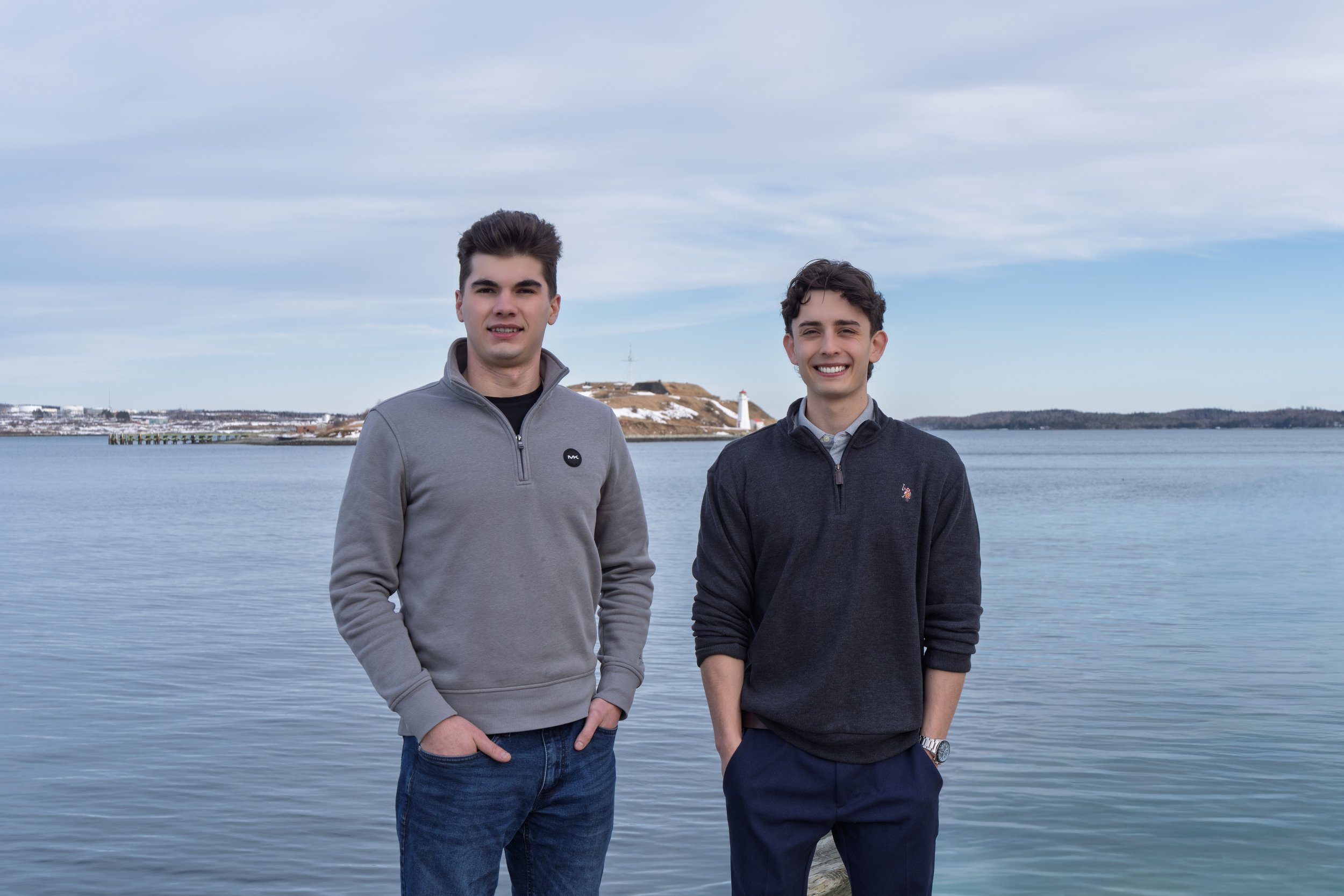 Two young men standing by a body of water with a hilly landscape, a lighthouse, and a cloudy sky in the background.