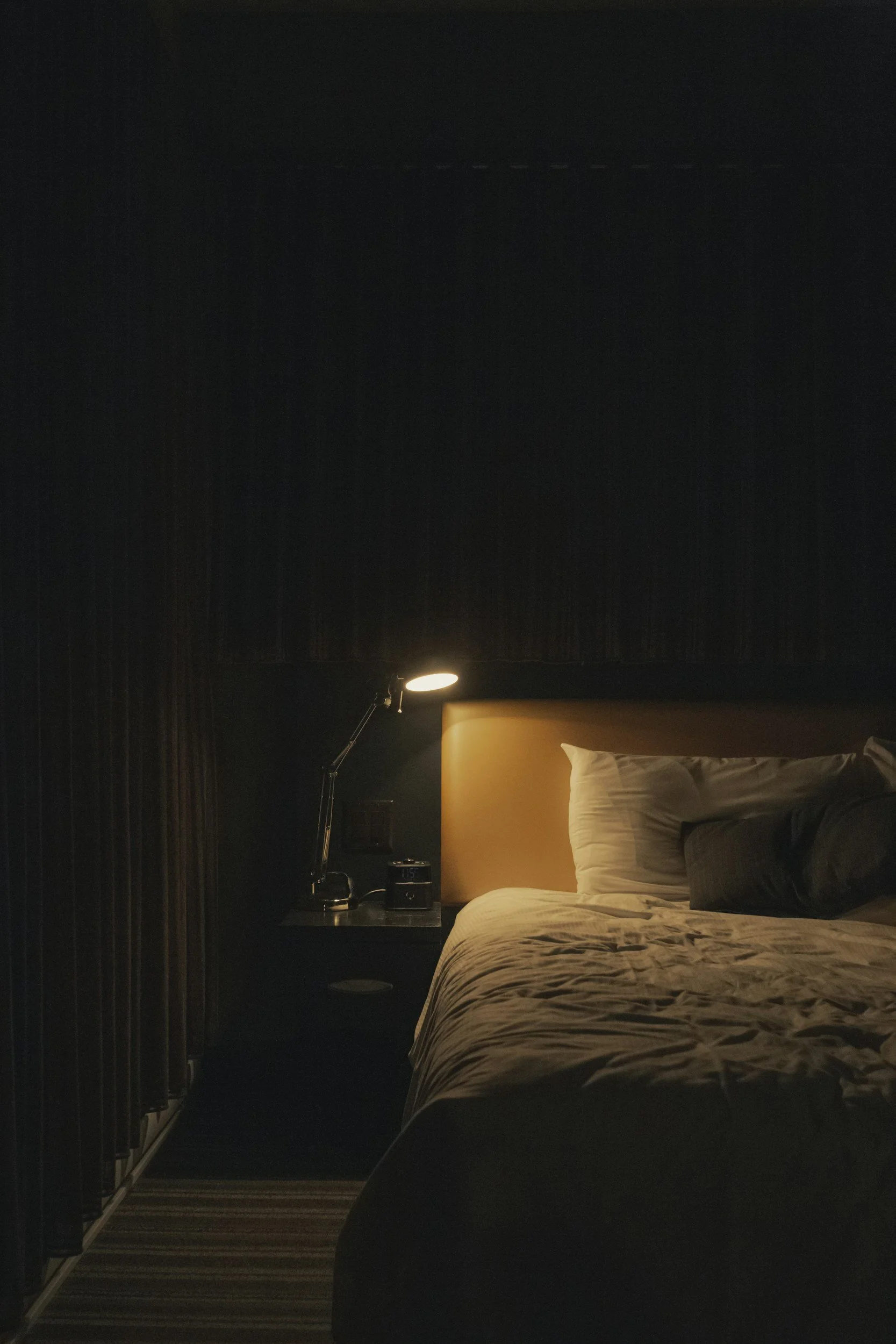 A dimly lit hotel bedroom with a bed, white pillows, black accent pillows, a bedside table with a lamp, clock, and other items, and black curtains.