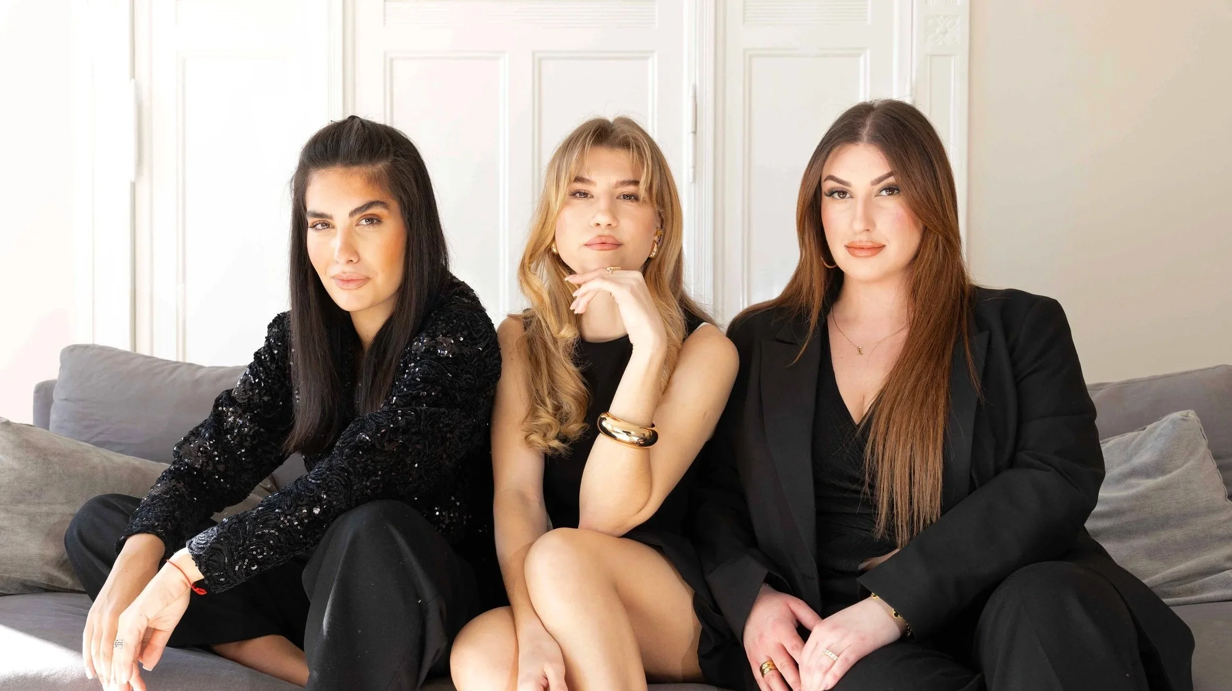 Three women sitting on a sofa in a room with white walls, dressed in black outfits, looking at the camera.