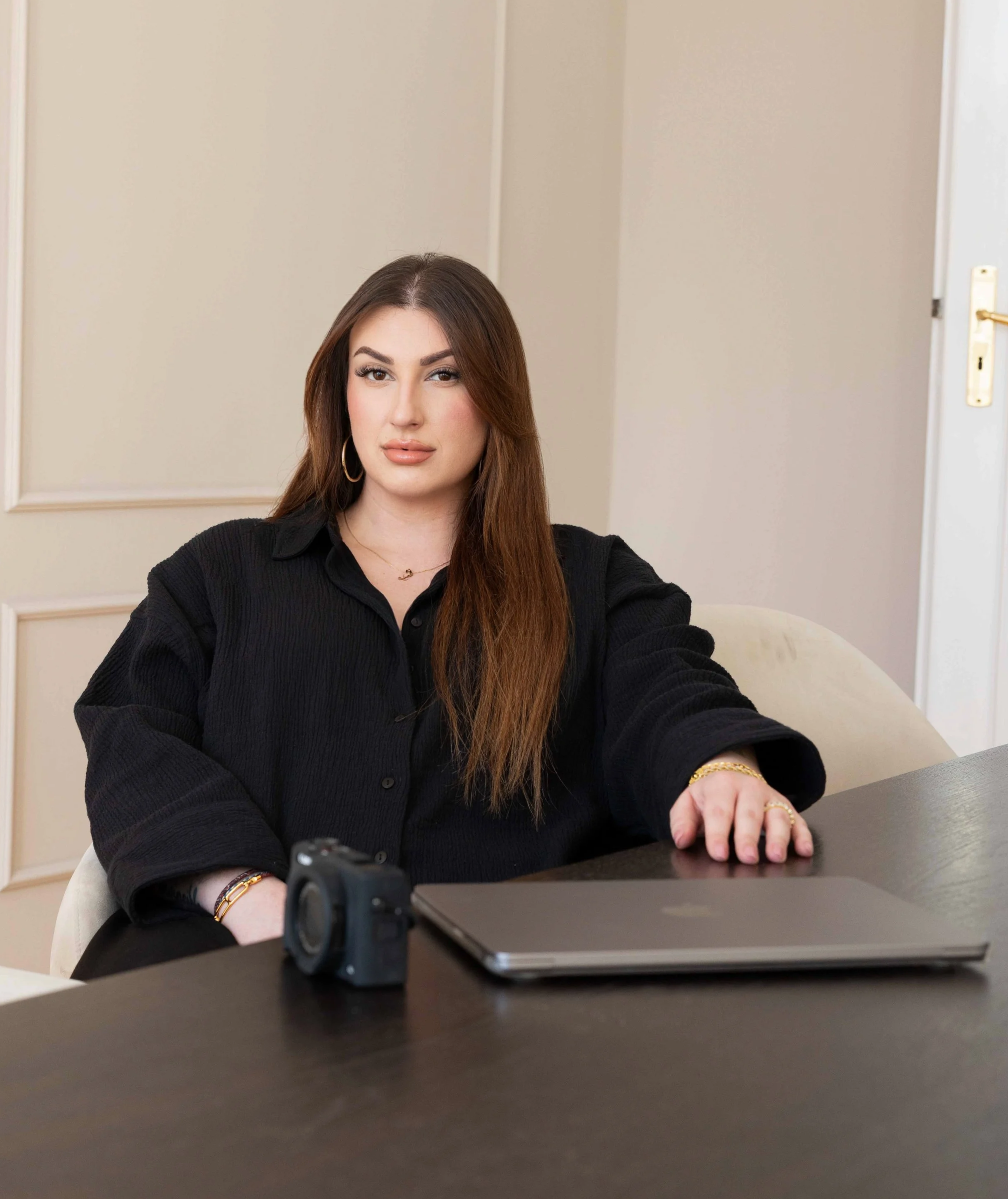 A woman with medium-length brown hair wearing a black shirt sitting at a table with a camera, a closed laptop, and gold jewelry.