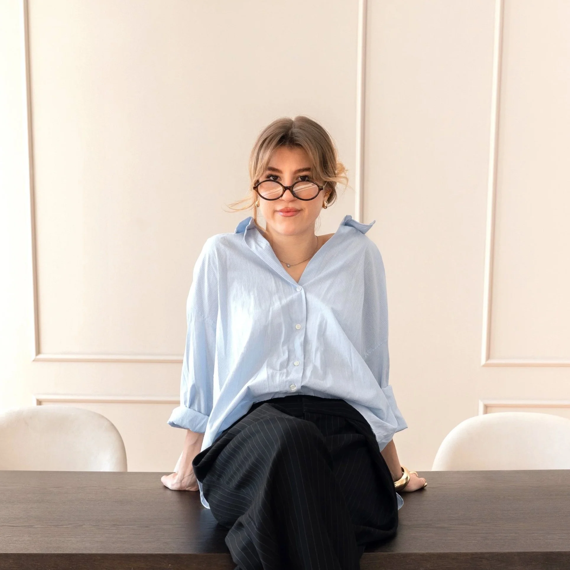 A woman with glasses and earrings sitting on a table, wearing a light blue shirt and black pants, in a minimalistic room with white walls.