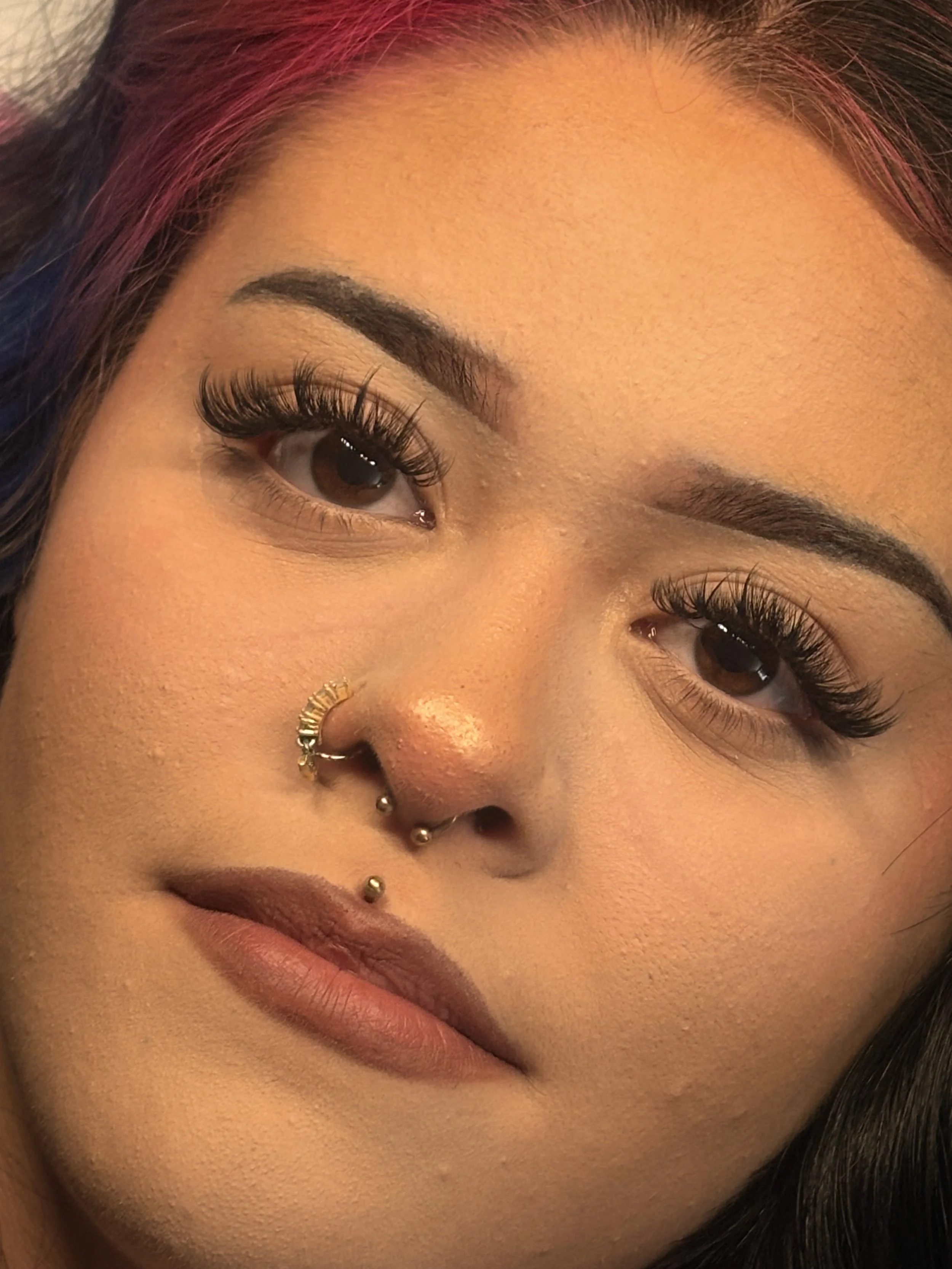 Close-up of a woman's face with makeup, including long eyelashes, eyebrow makeup, and multiple facial piercings in her nose and lips. Aura by Joogie real clients and real testimonials.