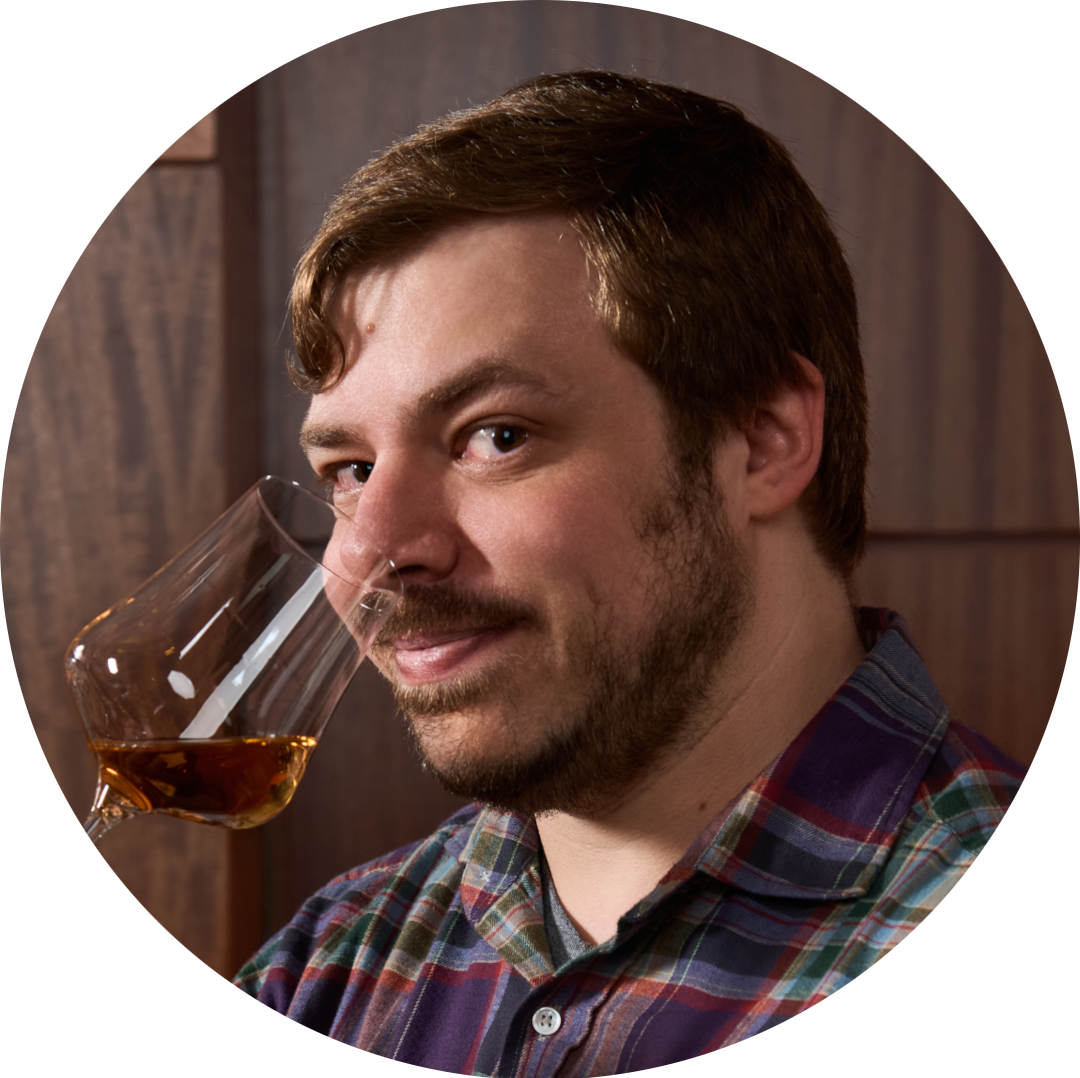 A man with brown hair and a beard holding a glass of amber-colored beverage near his face, smiling slightly, wearing a plaid shirt.