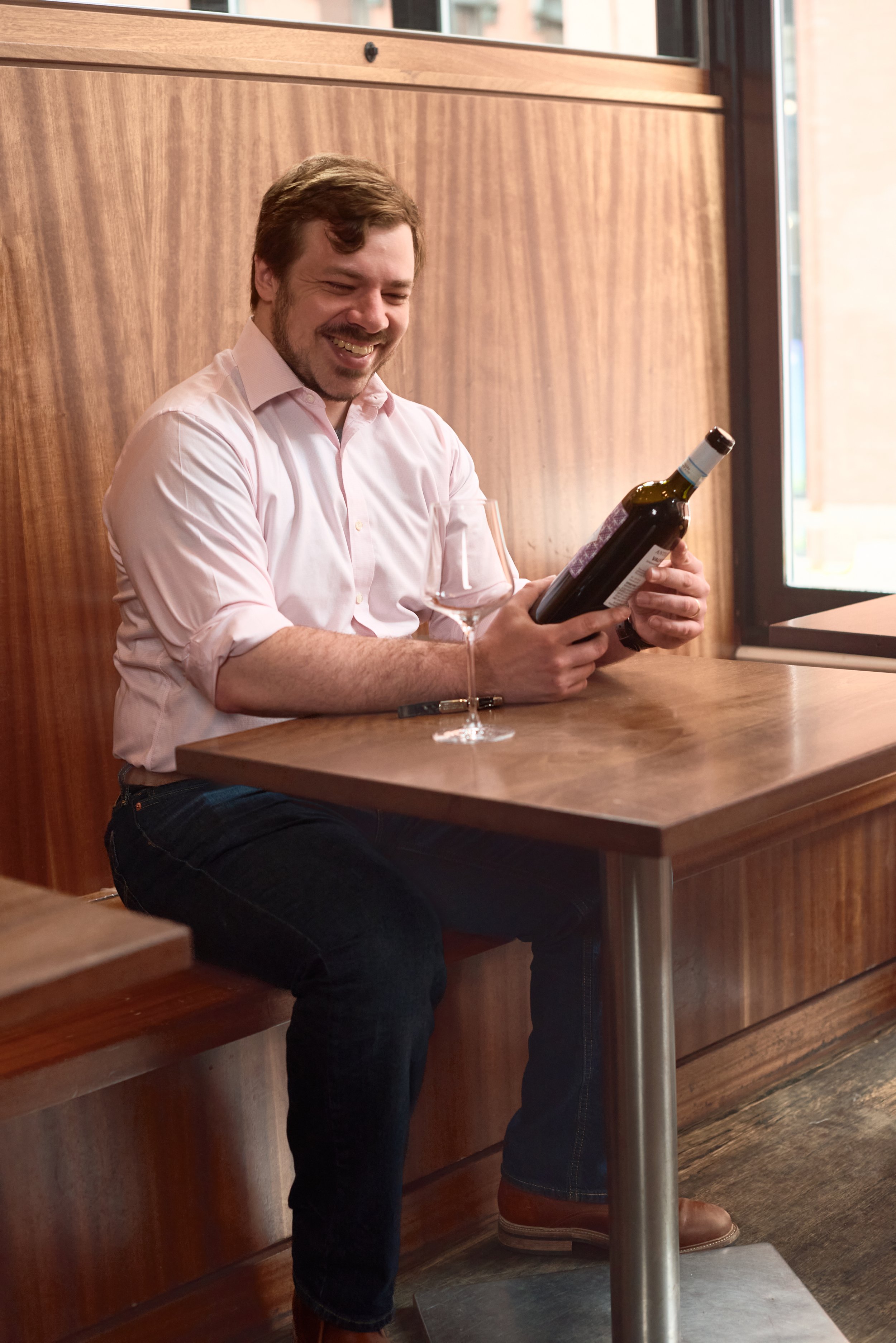 A man with brown hair and a beard, wearing a pink button-up shirt and dark jeans, sitting at a wooden table in a restaurant, holding a bottle of wine and smiling.