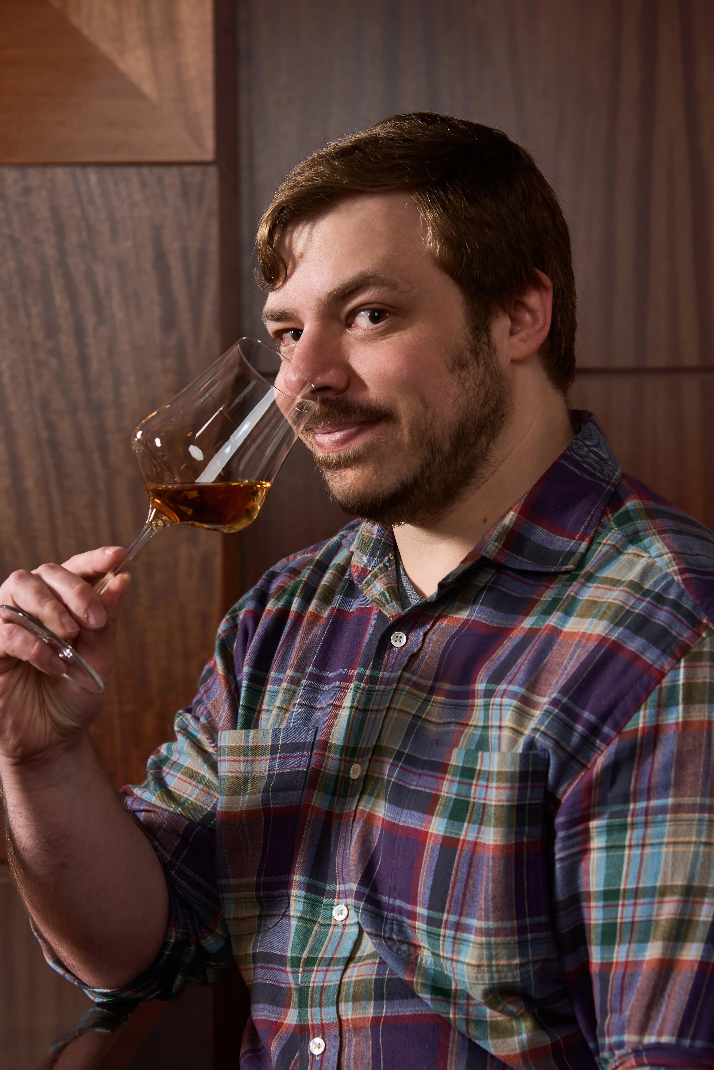 A man with a beard and brown hair, wearing a colorful plaid shirt, holds a glass of whiskey close to his face, standing against a wooden background.