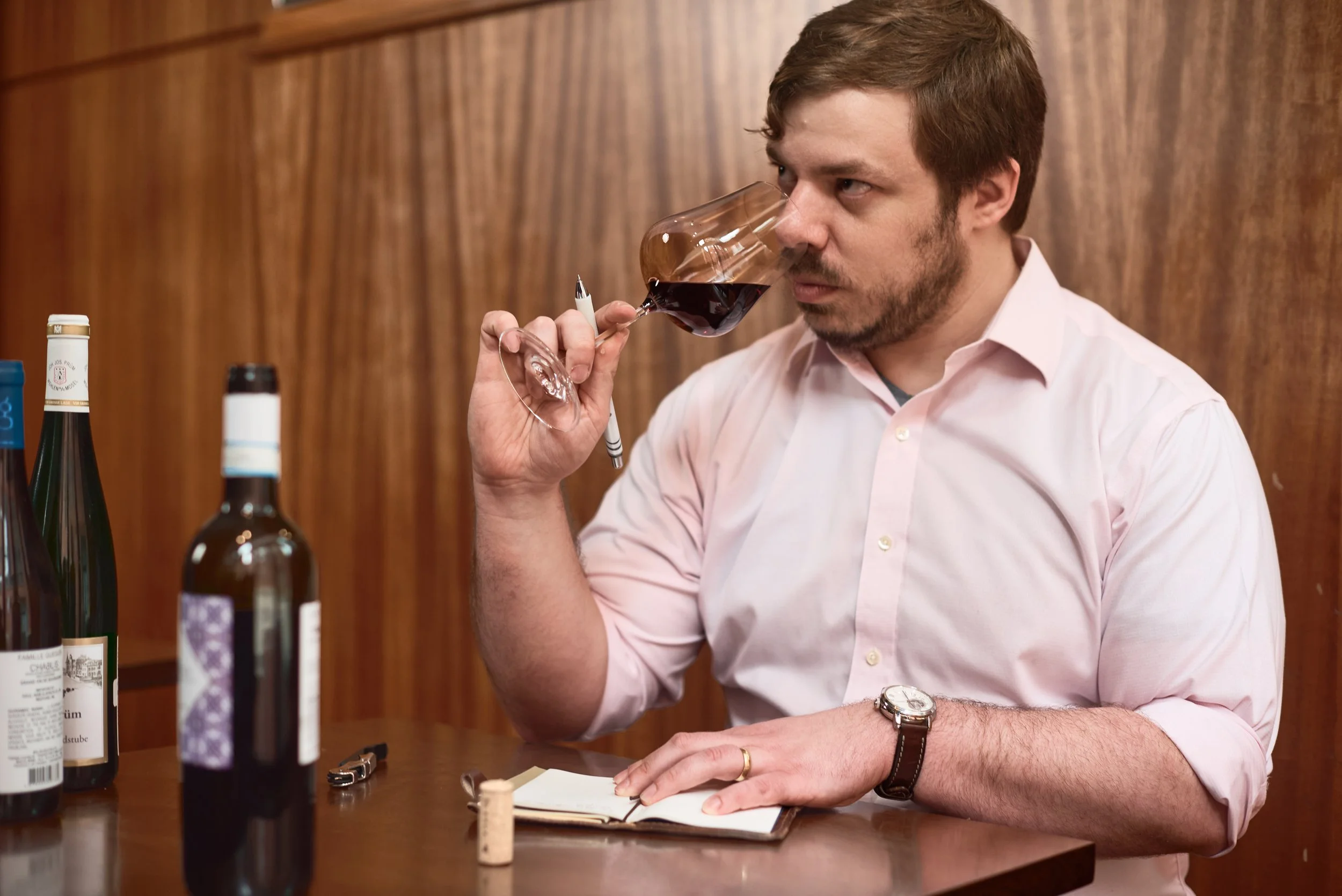 A man with light brown hair and a beard wearing a pink dress shirt is sitting at a table, smelling a glass of red wine. There are several wine bottles on the table and the background features a wooden wall.