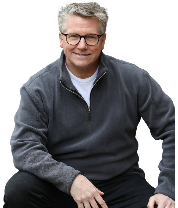 A middle-aged man with gray hair and glasses, smiling, wearing a gray zippered jacket over a white t-shirt, sitting with a relaxed posture.