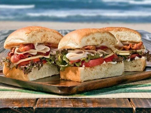 Three submarine sandwiches with turkey, ham, lettuce, tomato, and cheese on a wooden tray by the beach.