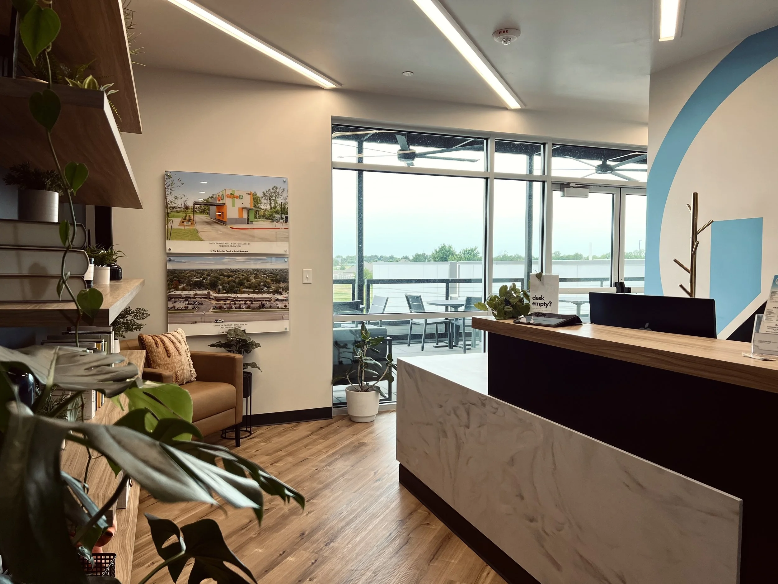 Modern office reception area with wooden flooring, a reception desk with marble panel, potted plants, a brown armchair with pillows, wall art, and a balcony with outdoor seating and a view of trees.