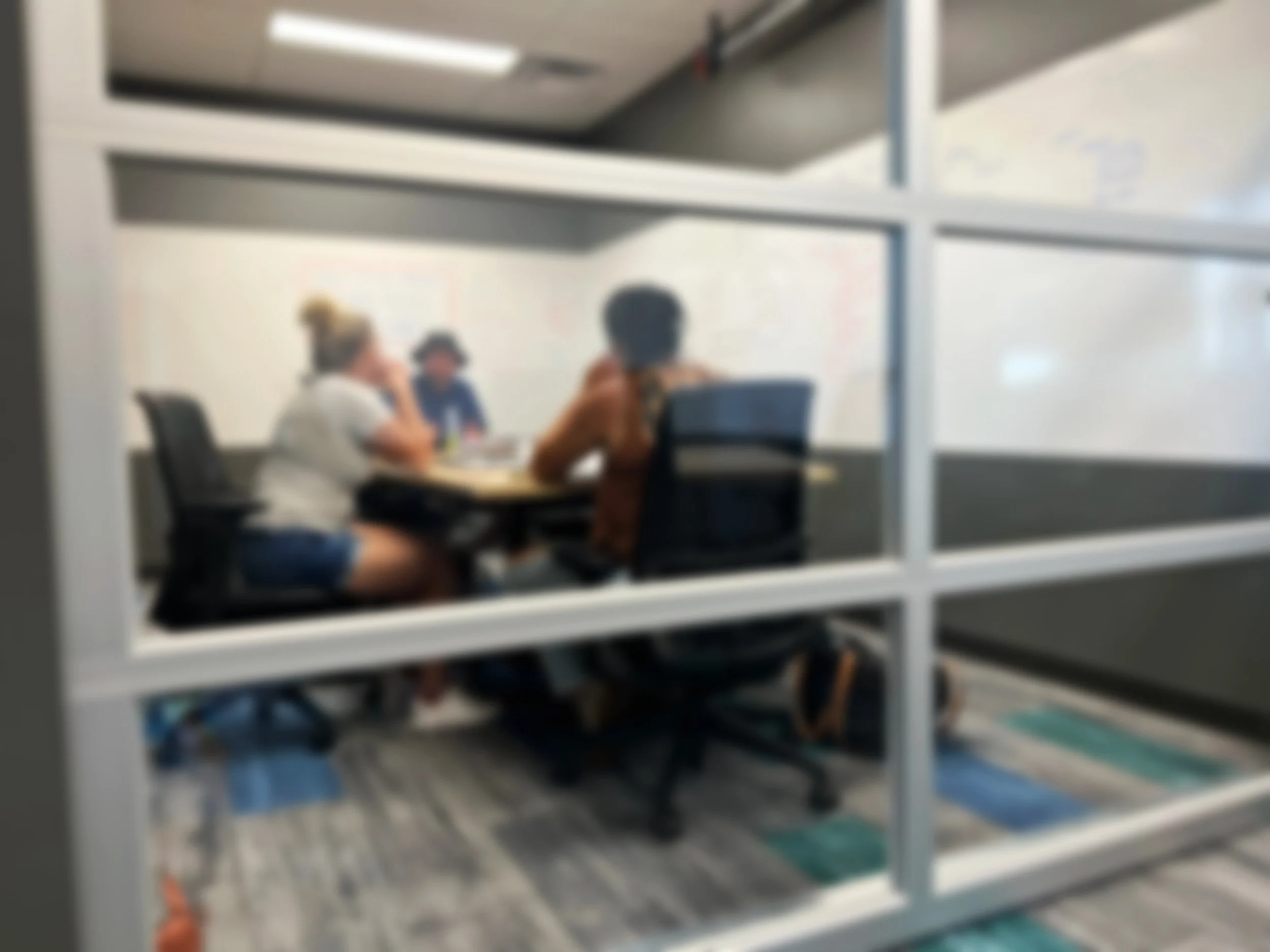 Three people sitting around a table in a small conference room, viewed through a glass panel.