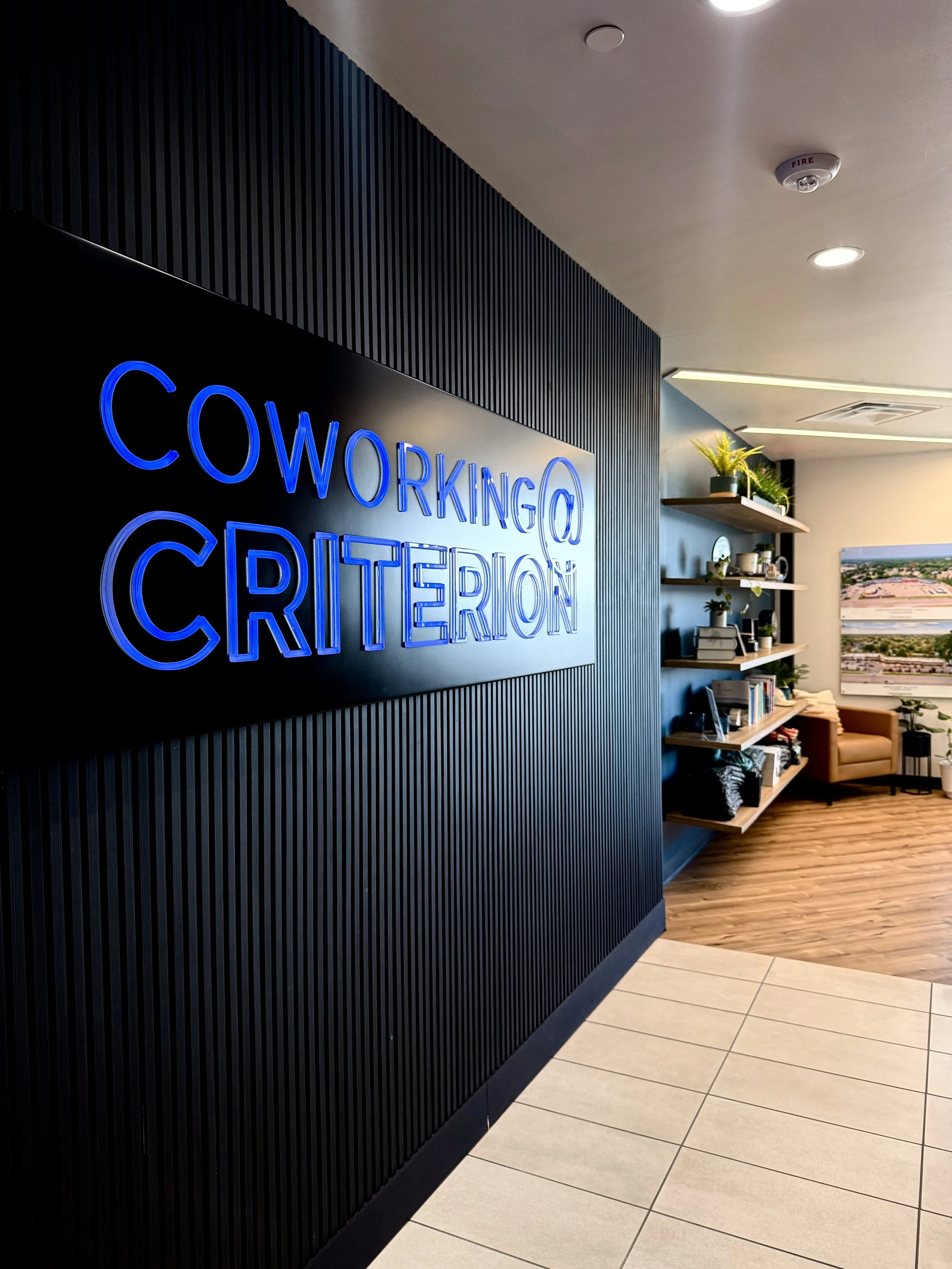 Interior of an office with a neon sign reading 'Coworking @ Criterion' mounted on a black, ribbed wall, with a bookshelf and seating area visible in the background.