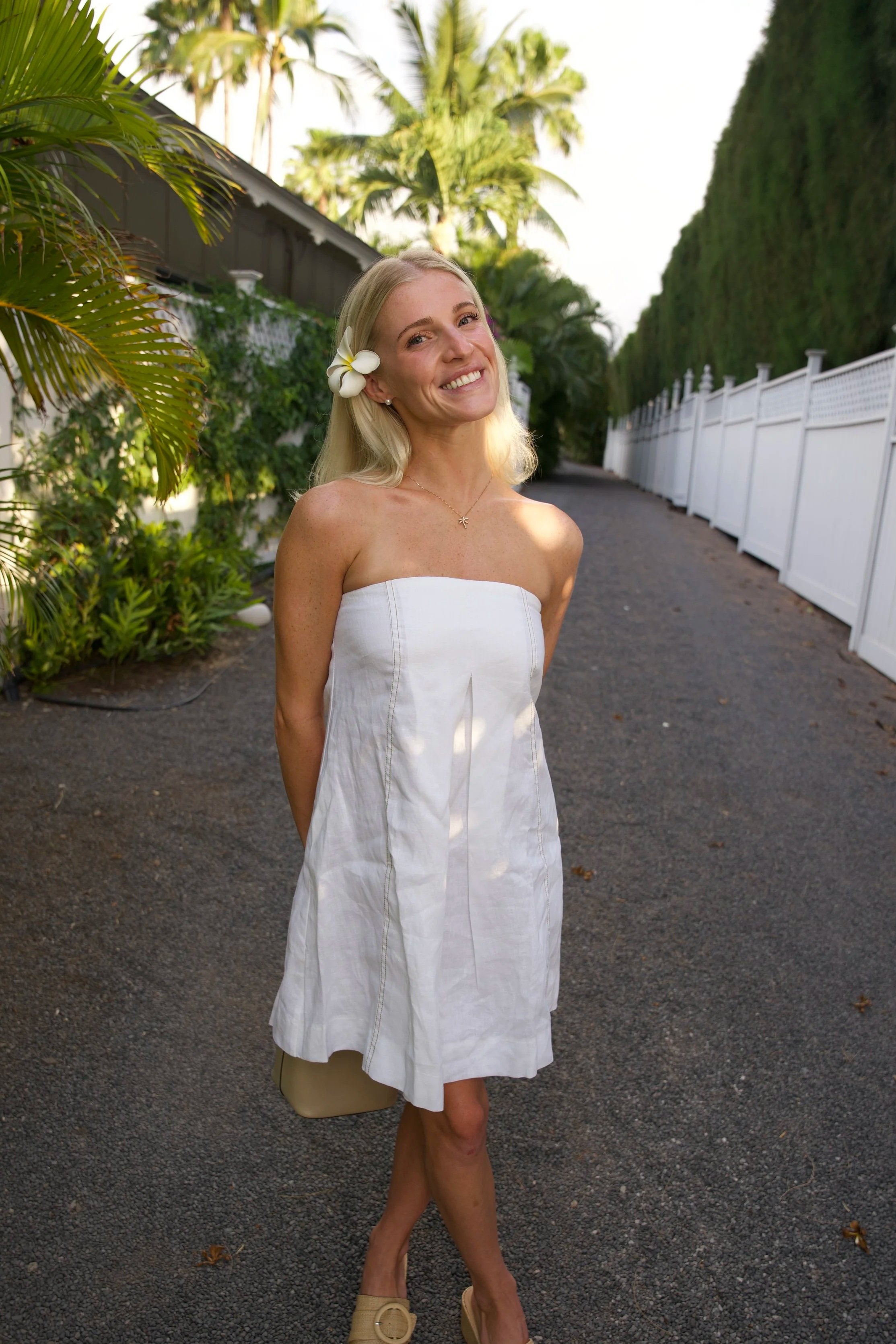 A smiling woman with long blonde hair, wearing a white strapless dress with a flower in her hair, stands on a paved path surrounded by lush tropical plants and tall trees. A white fence runs along the side of the path.