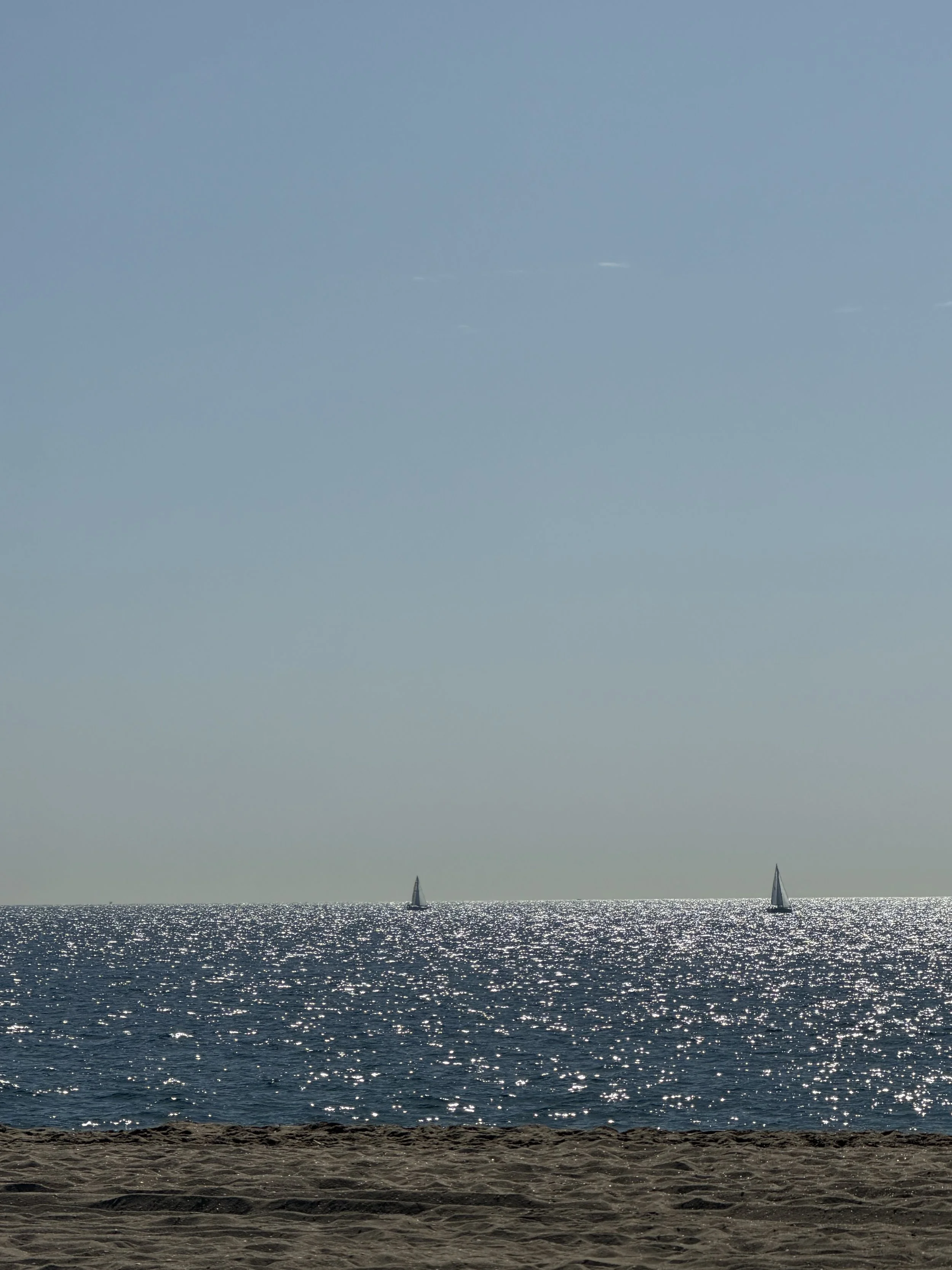 Sailboats on the ocean near a sandy beach with sunlight reflecting off the water, under a clear sky.