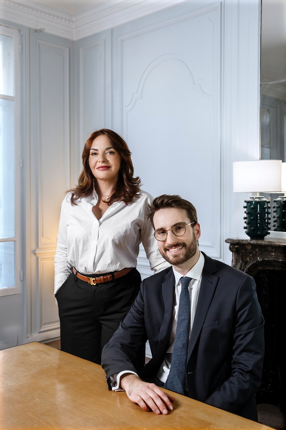 Une femme debout à côté d'un homme assis devant un bureau dans une pièce élégante avec des murs en moldura et un feu de cheminée en marbre, tous deux habillés de manière professionnelle. La femme porte une chemise blanche et un pantalon noir, l'homme porte un costume sombre avec une cravate."