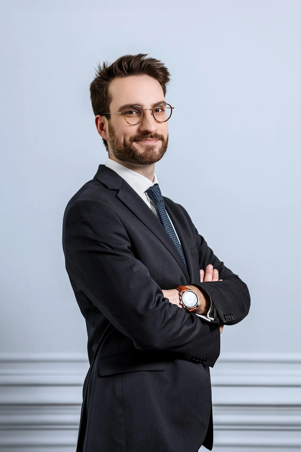 Un homme d'affaires portant un costume noir, des lunettes rondes, une montre en cuir et une cravate, posant avec les bras croisés contre un mur clair.
