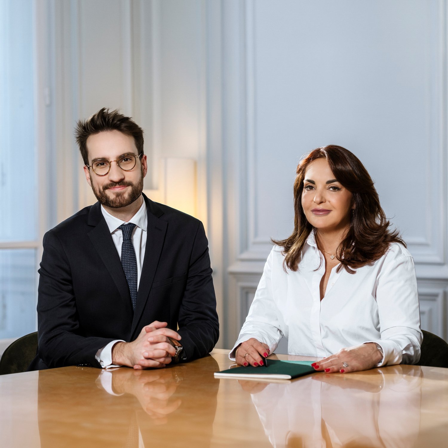 Deux personnes assises à une table en bois clair dans une pièce aux murs blancs et moulurés, avec une lampe d'appoint à l'arrière. L'homme porte un costume noir, une chemise blanche, une cravate à pois et des lunettes. La femme porte une chemise blanche et a les cheveux bruns ondulés.
