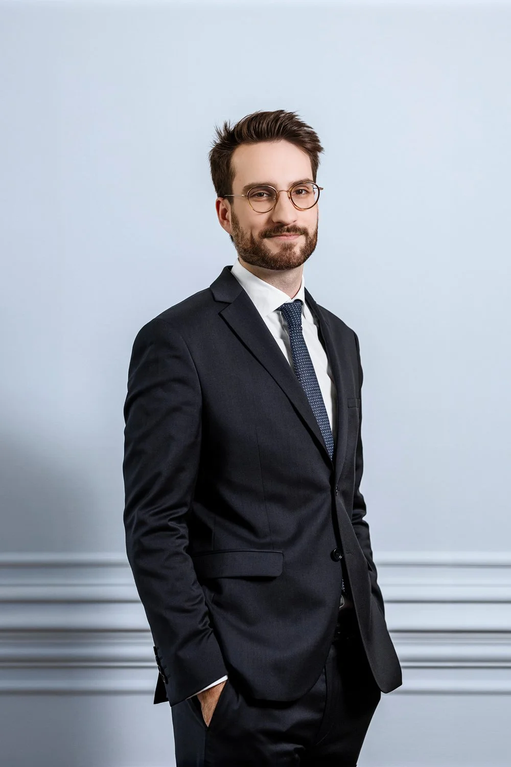 Portrait d'un homme portant un costume sombre, chemise blanche, nœud papillon, lunettes rondes, foncé avec des cheveux et une barbe bruns, debout contre un mur gris clair.