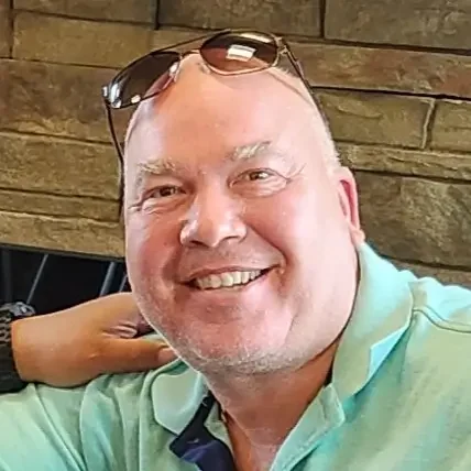 Smiling man with sunglasses on his head, wearing a light green shirt, in front of a wooden background.