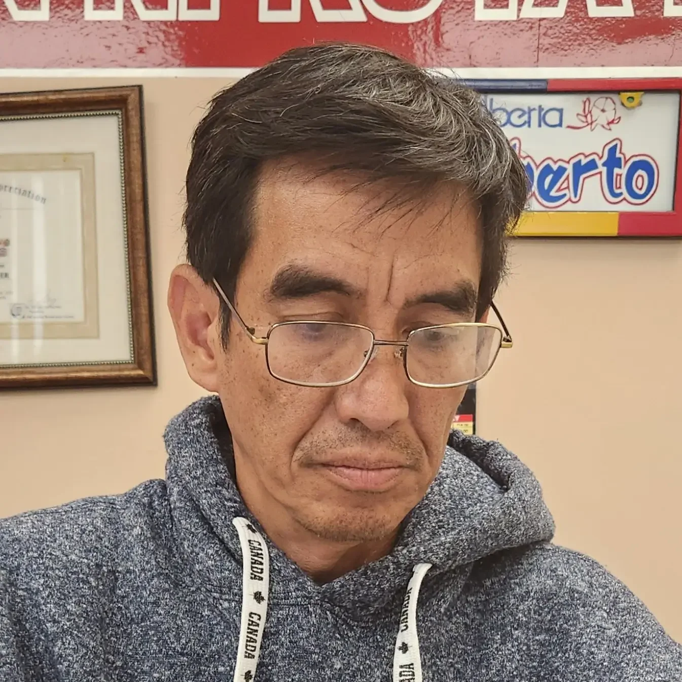 A man with glasses and a gray hoodie looking down in an indoor setting with framed pictures on the wall behind him.