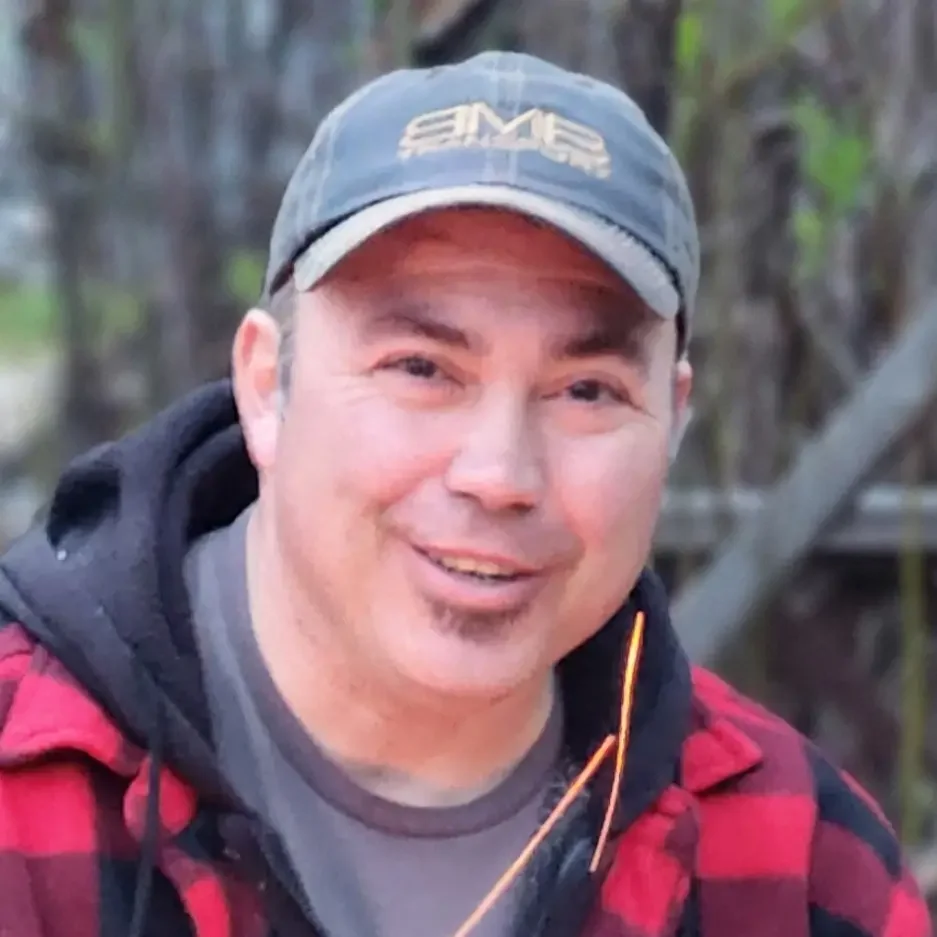 A smiling man wearing a gray cap with a logo, a black hoodie under a red and black plaid jacket, outdoors with trees in the background.