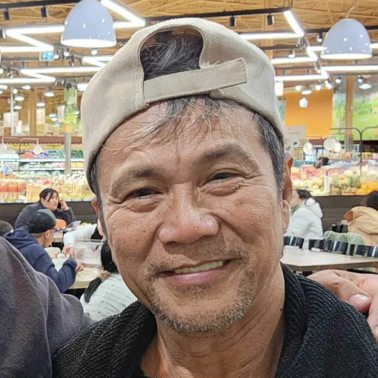 A smiling middle-aged man wearing a beige cap backward in a busy grocery store with people shopping and fresh produce in the background.