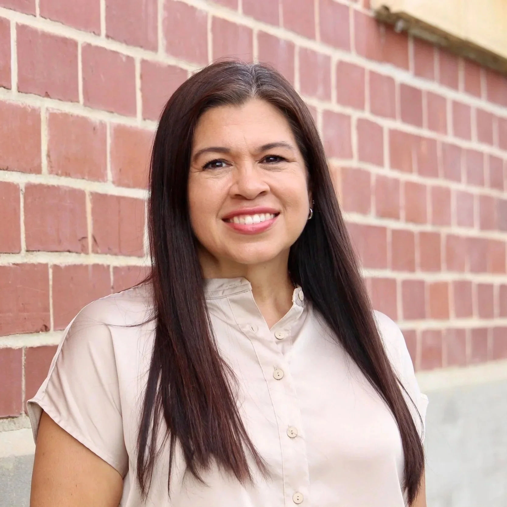 A woman with long dark hair standing outdoors in front of a red brick wall, smiling at the camera.