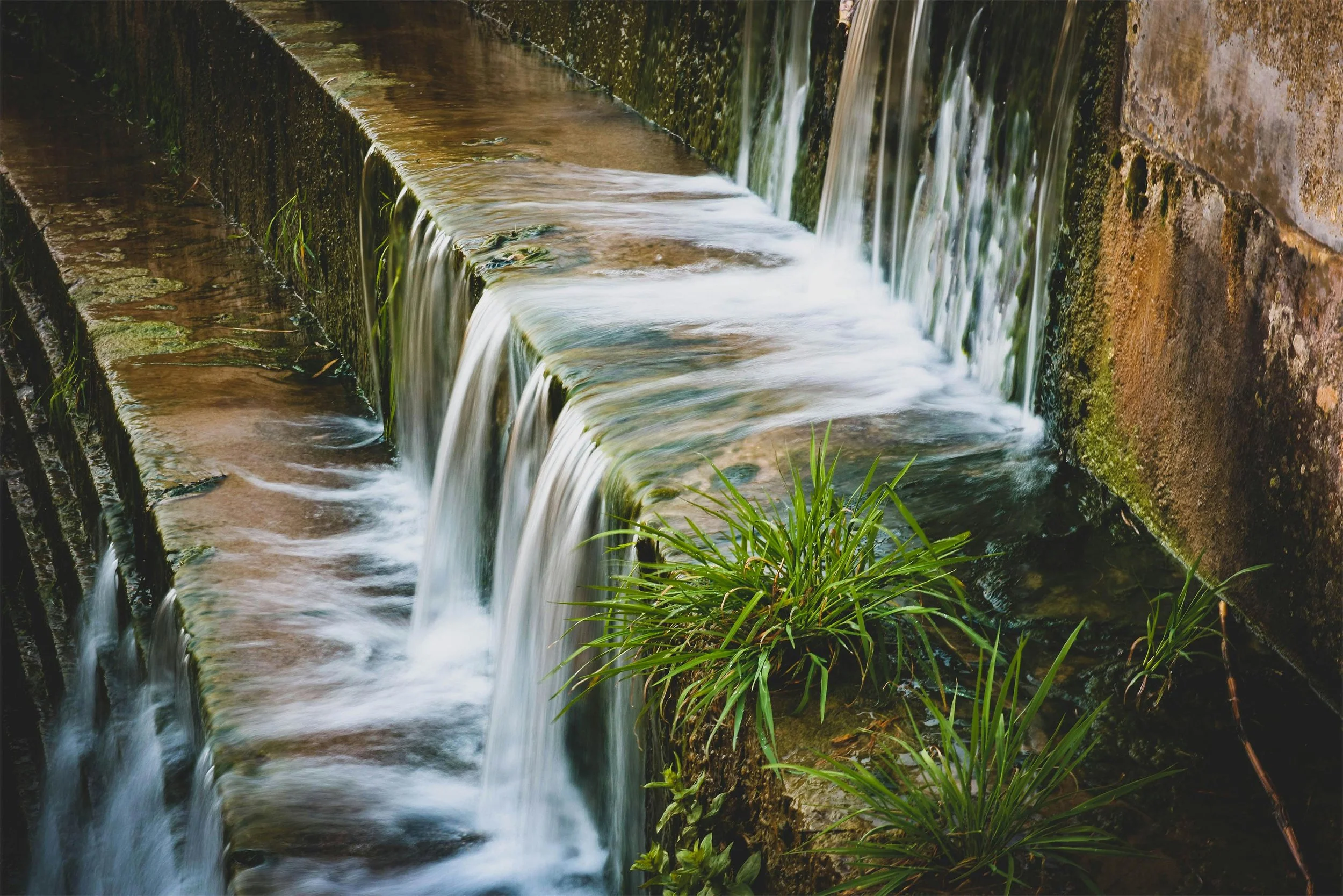 Small waterfall or creek flowing over mossy, weathered brick or concrete structure, with green grass and plants growing at the edge.