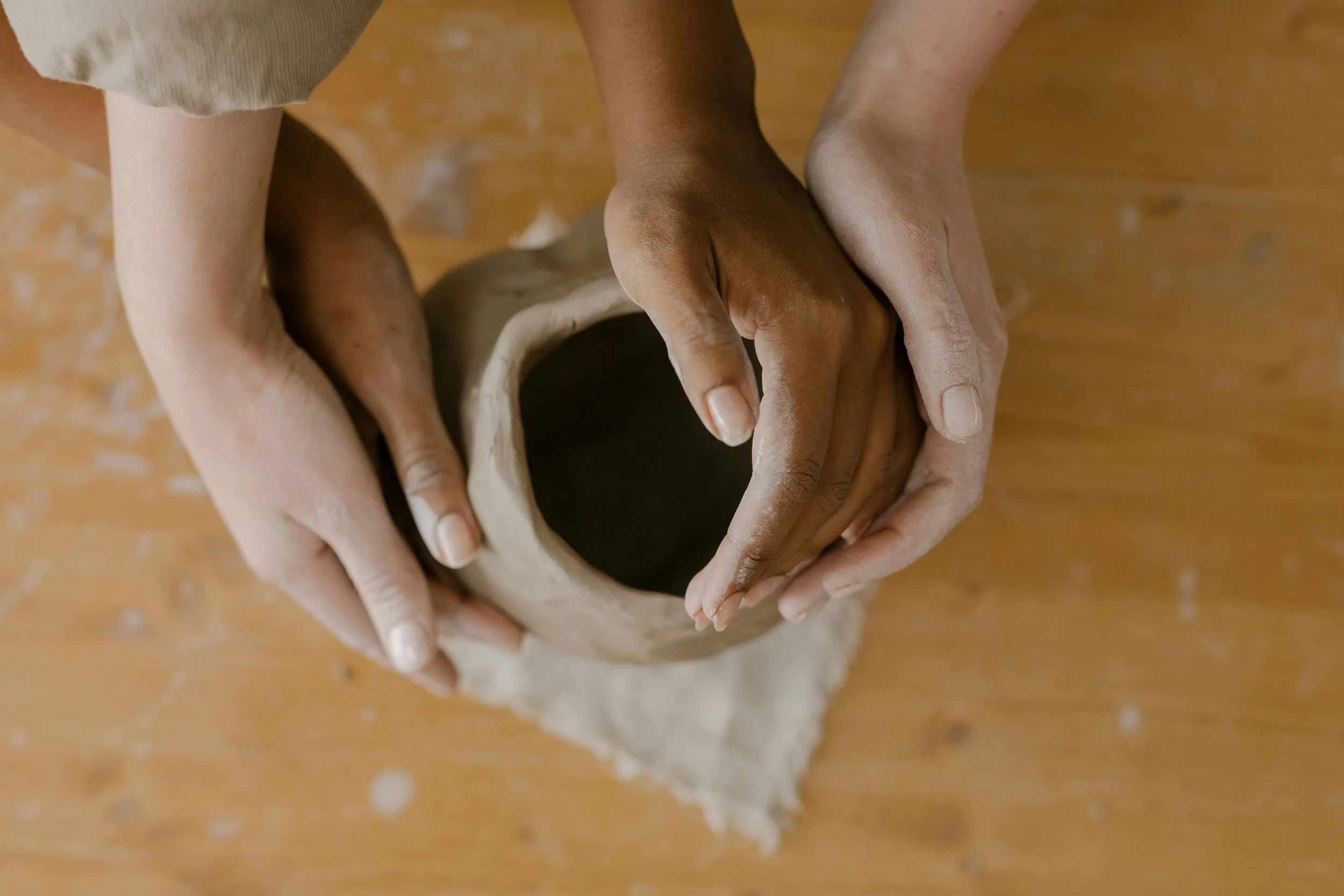Two people crafting a small clay pot together on a wooden surface, with one person's hands guiding the process.