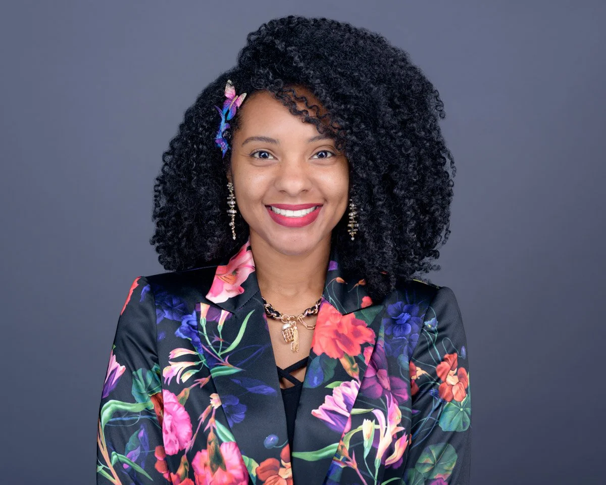Portrait of a woman with curly black hair wearing a colorful floral blazer and jewelry, smiling against a gray background.