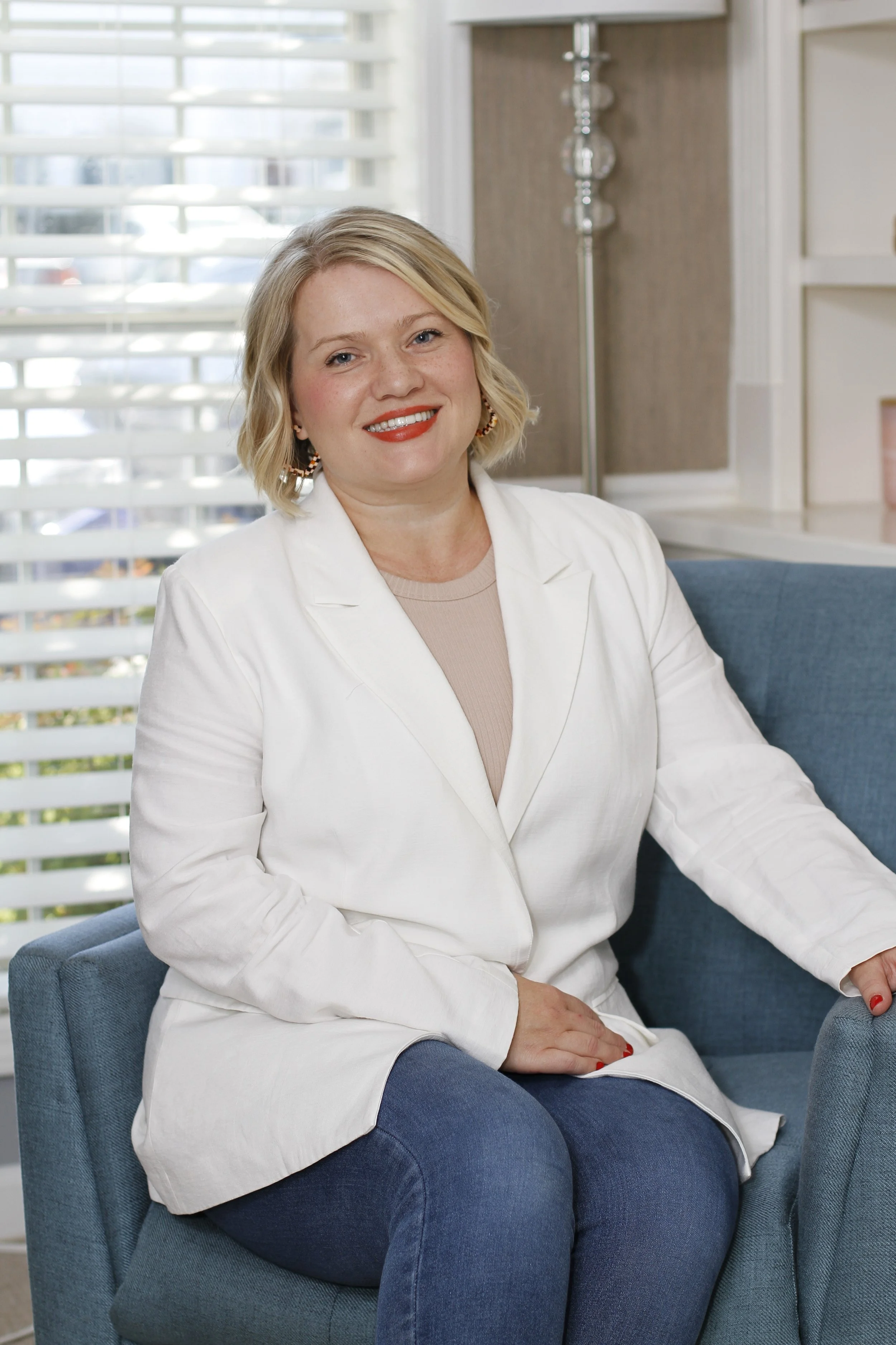 A woman with blonde hair seated on a blue armchair in a bright room with white blinds, wearing a white blazer and jeans.