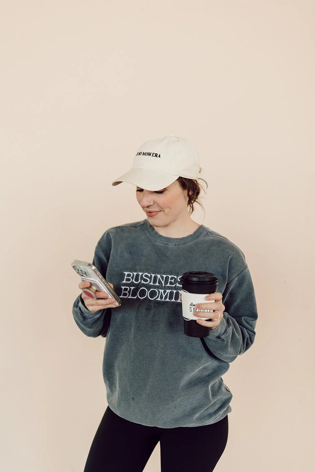 Woman in gray sweatshirt and white baseball cap holding smartphone and coffee cup, standing against plain background.