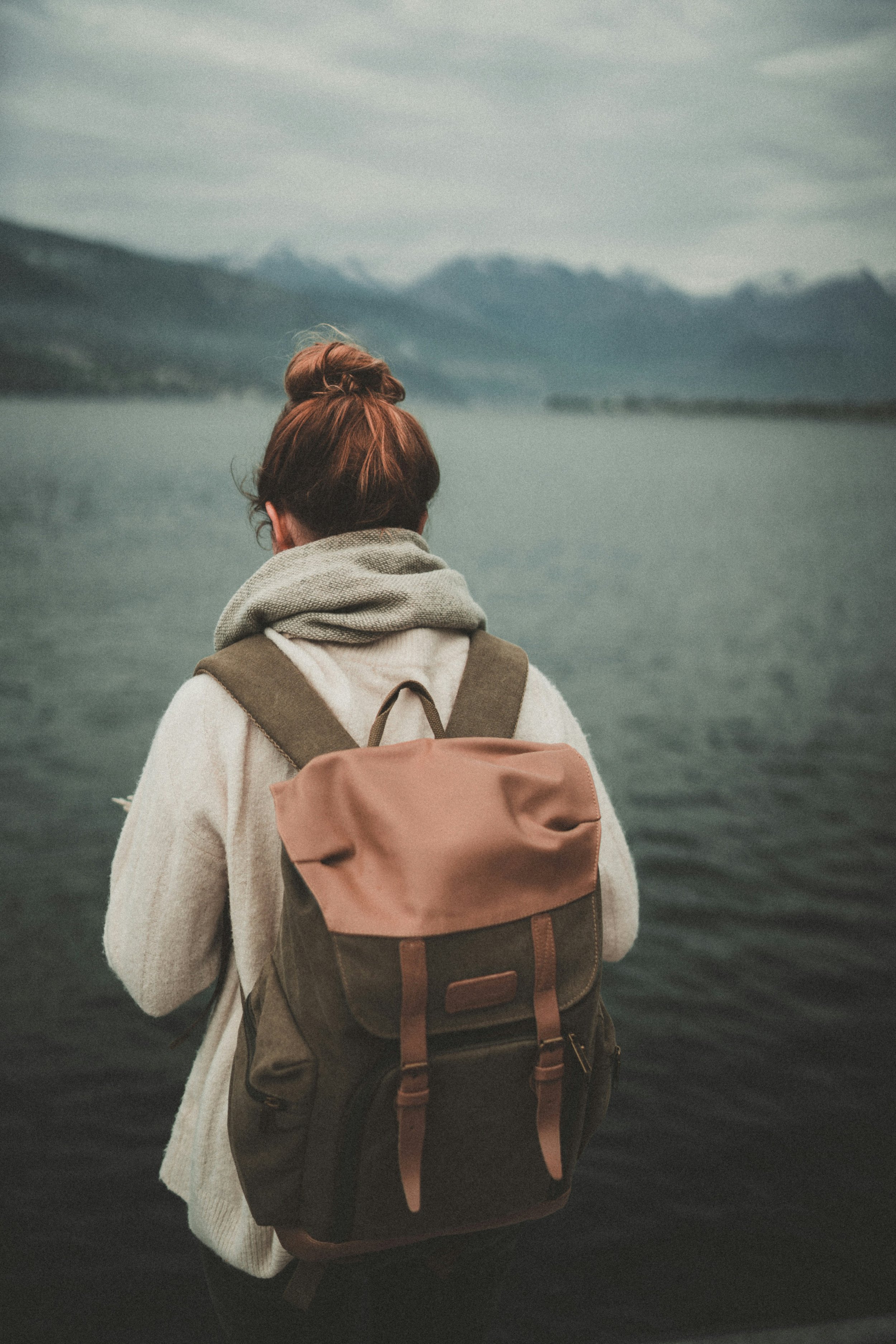 A person with reddish-brown hair tied in a bun, wearing a beige sweater, gray scarf, and carrying a brown and green backpack, is standing by a large body of water with mountains in the background during overcast weather.