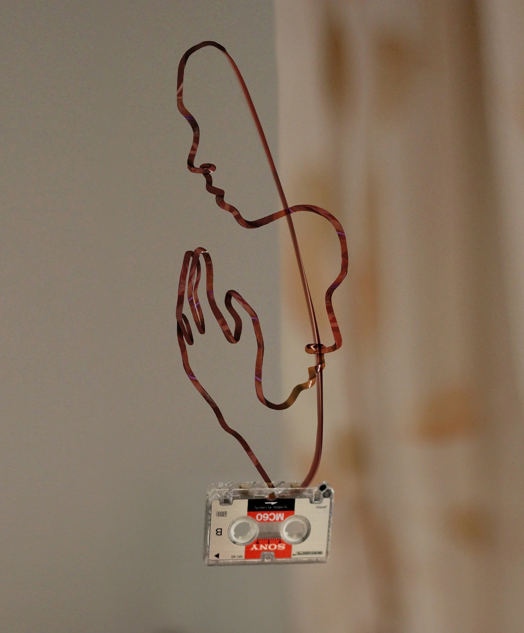 Wall-mounted art piece resembling a stylized human face and hand, made of bent copper wire, with a cassette tape at the base.