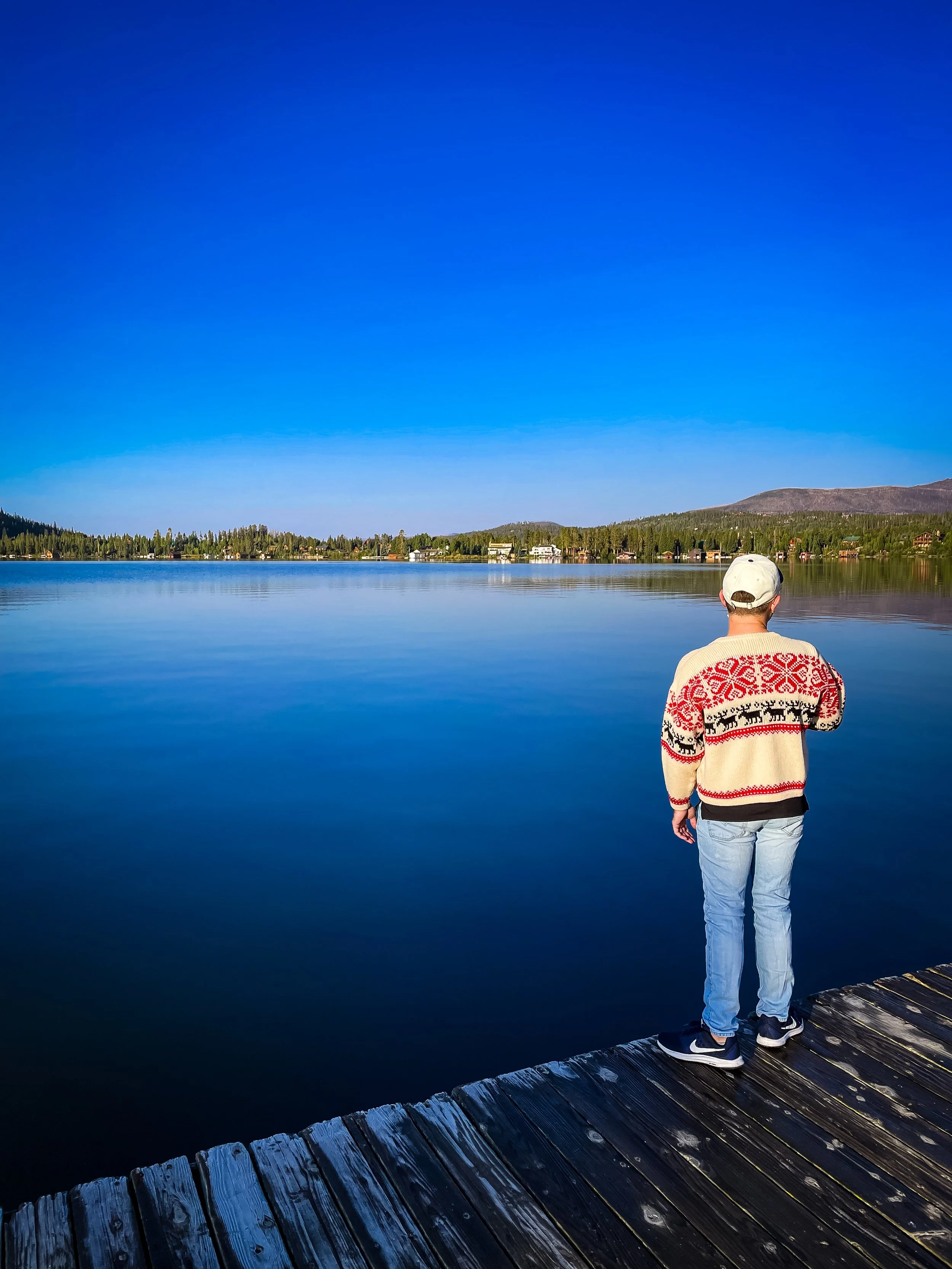 Person standing on a wooden dock overlooking a calm lake with houses and mountains in the distance under a clear blue sky.