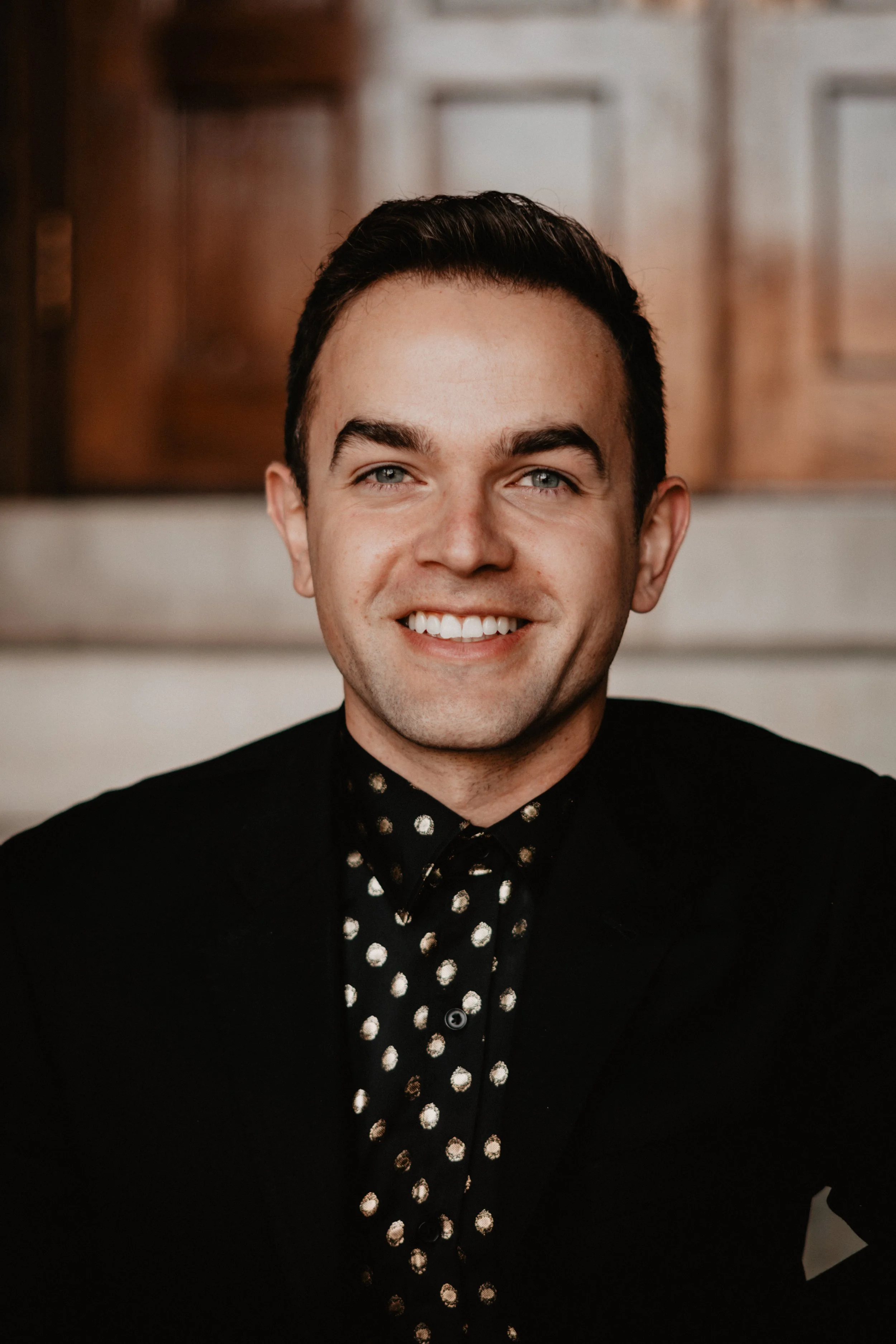 A young man with short dark hair, blue eyes, and a bright smile, wearing a black blazer and a black shirt with white polka dots, sitting indoors with a blurred wooden background.
