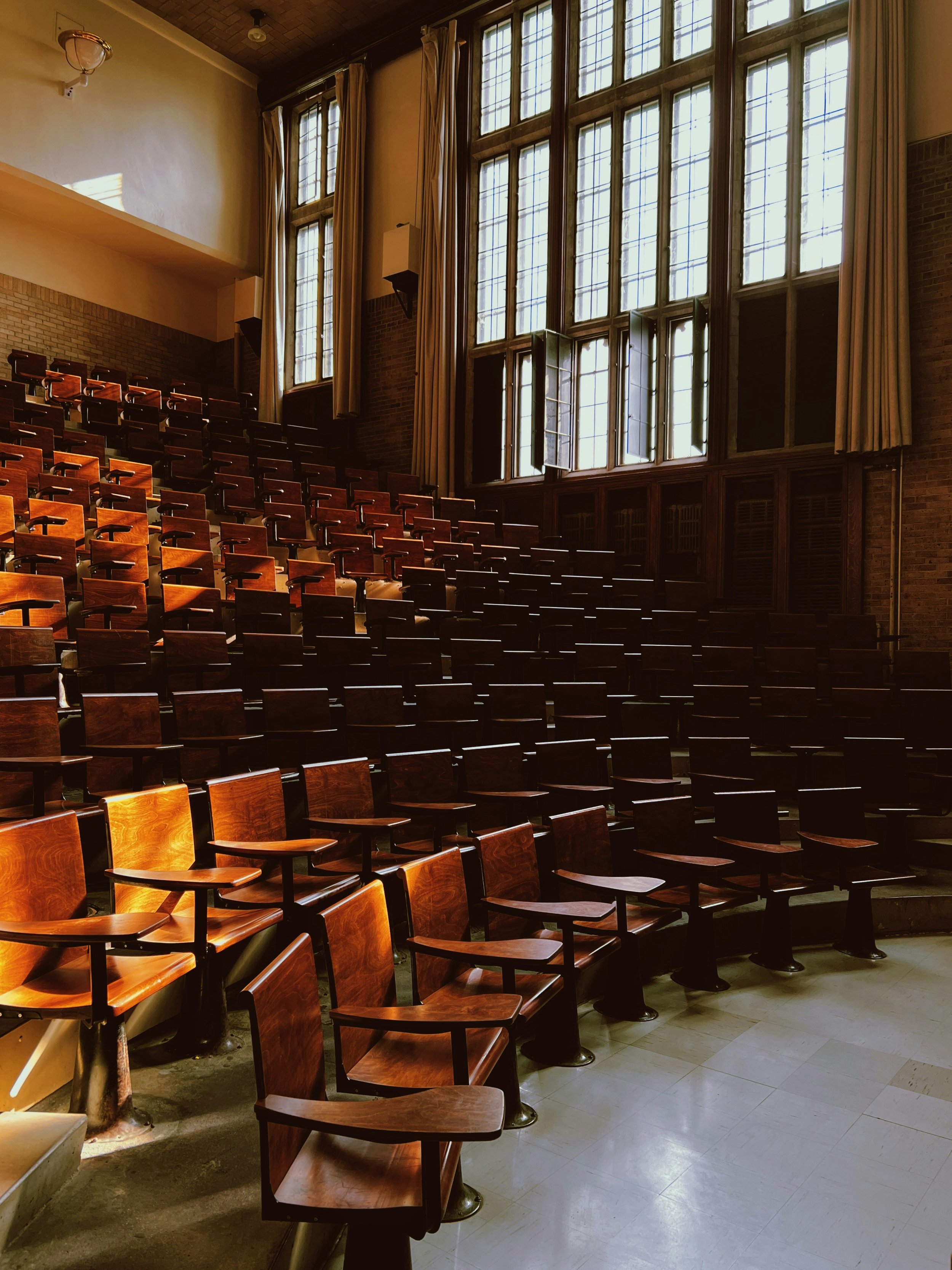 Empty lecture hall with rows of wooden seats and large windows letting in sunlight.