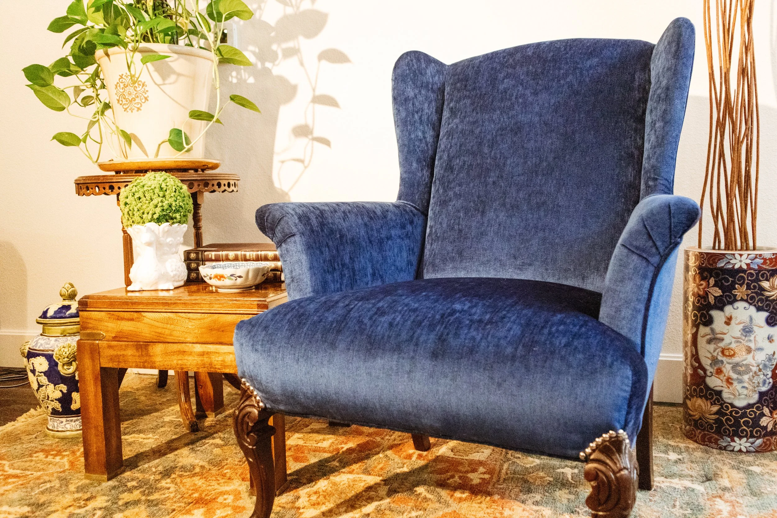 A blue upholstered armchair with carved wooden legs. Next to it is a wooden side table holding a white vase with green plants, a small decorative bowl, and a box of books. To the side, there are additional decorative vases and a tall bundle of dried branches. The setting is a cozy interior with a patterned rug on the floor.
