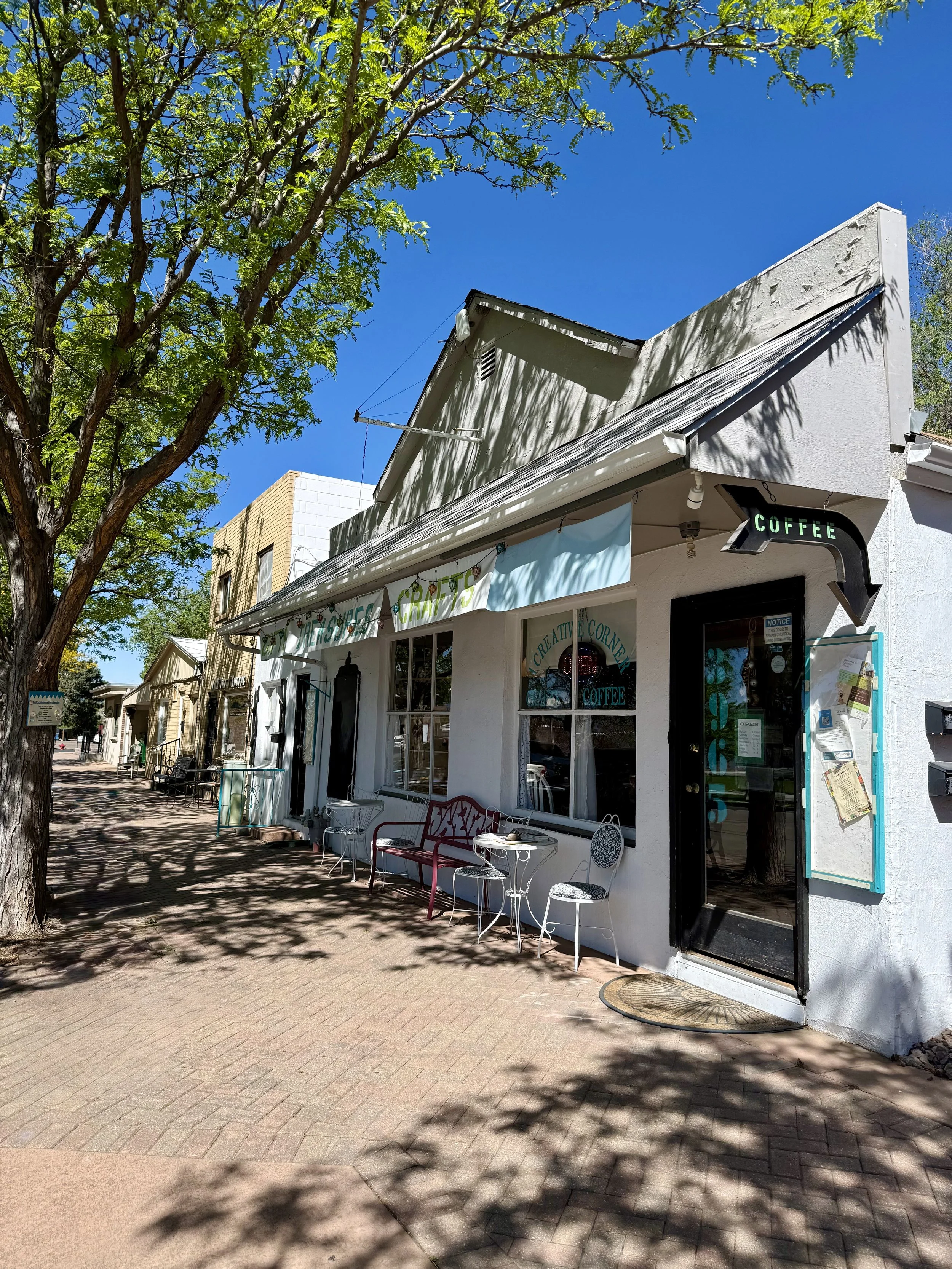 A quaint outdoor cafe storefront, The Creative Corner, on a sunny day with white walls, black door, and seating outside. A green sign above the door reads 'COFFEE'. The sidewalk is shaded by a large leafy tree. The Coffee Shop is in Westminster, CO.
