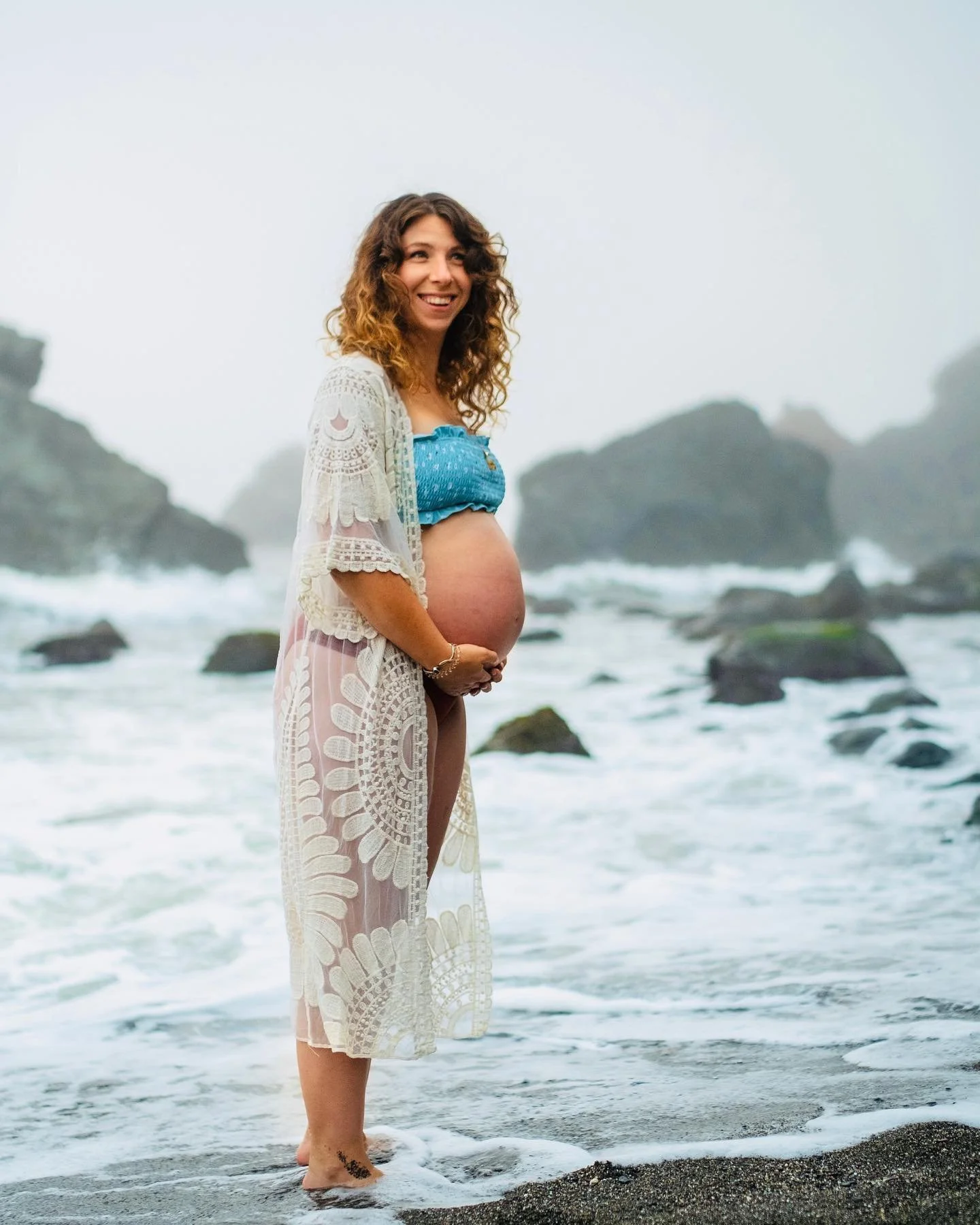 A certified doula near me in Massachusetts, Courtney stands in the ocean waves holding her pregnant belly with a smile.