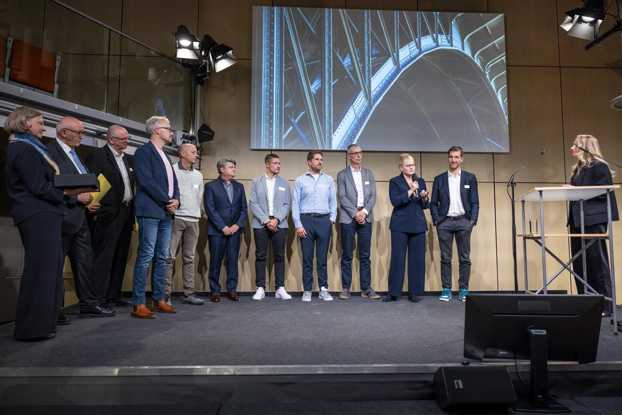 Das Team des Chemnitzer Viadukts mit Urkunden auf der Bühne nach der Verleihung des Staatspreises, rechts die Moderatorin am Pult